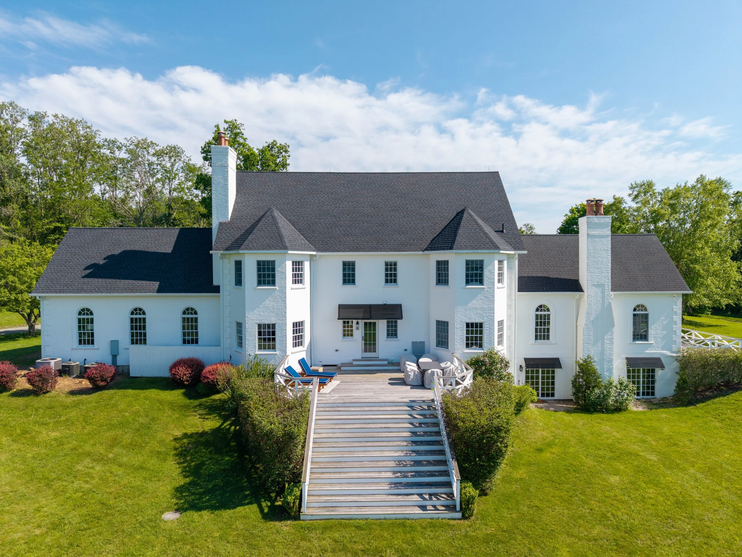 A large white two-story house with black shingles and multiple chimneys, surrounded by green grass and trees, with a wide staircase leading to the back entrance and an outdoor deck with lounge chairs.
