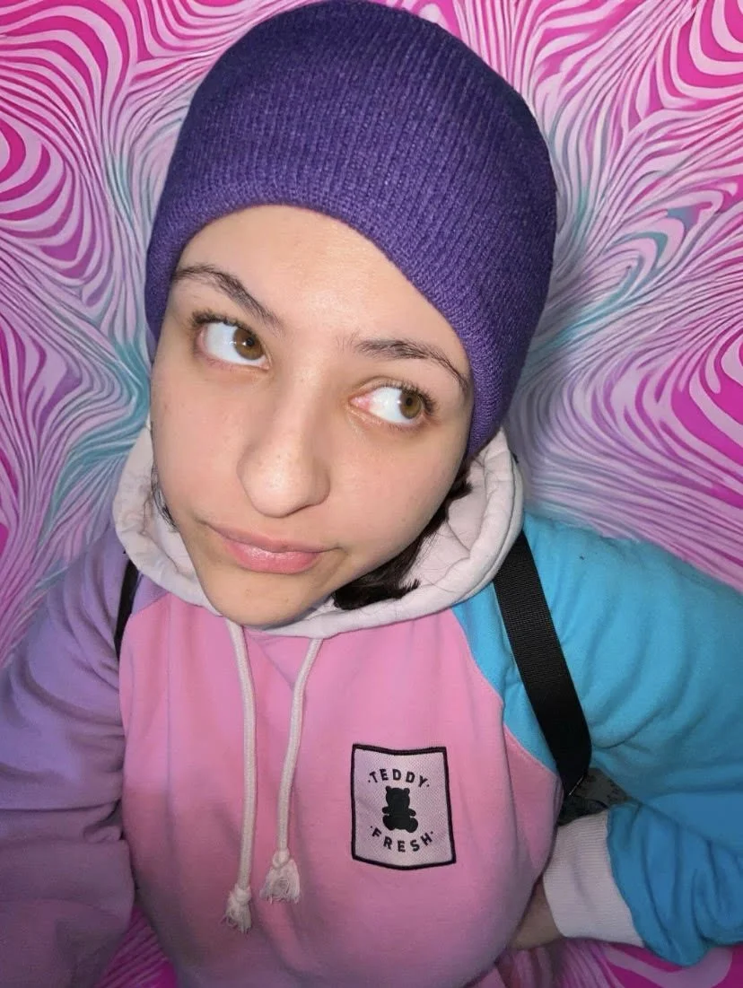 A person wearing a purple beanie and a pastel-colored hoodie, lying on a pink and blue marbled background, looking upward with a thoughtful expression.