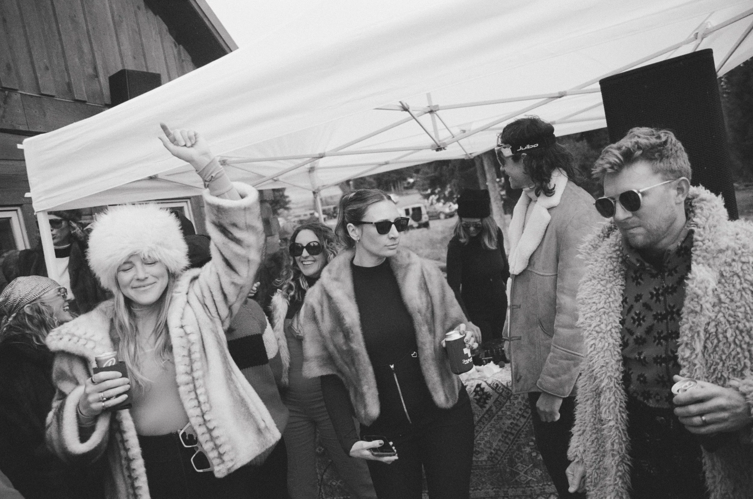 Group of people outside under a canopy, wearing winter clothing, some holding drinks, with trees and parked cars in the background.