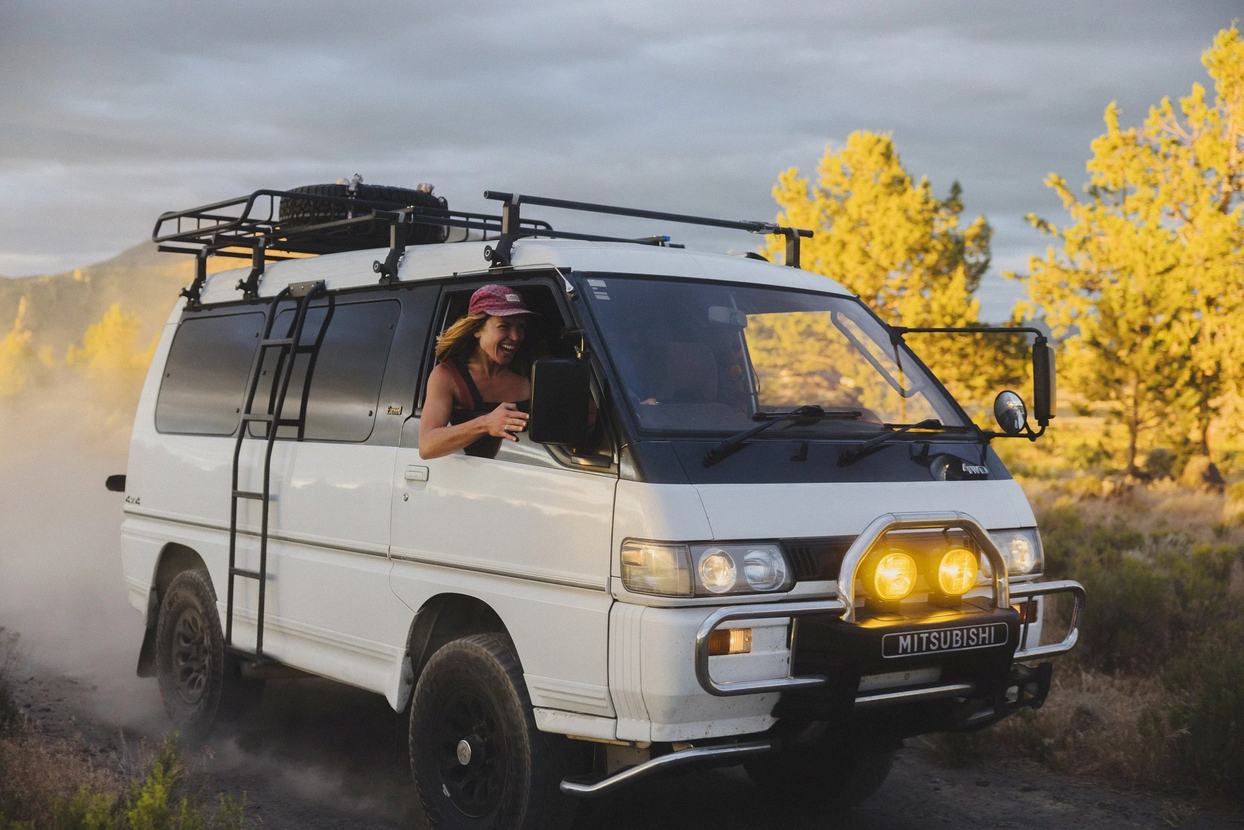 A woman with a hat is sitting in the driver's seat of a white Mitsubishi off-road vehicle, which is kicking up dust as it drives on a dirt road. The vehicle has a roof rack, side ladder, and front bumper lights. In the background, there are yellow-leaved trees and a cloudy sky.