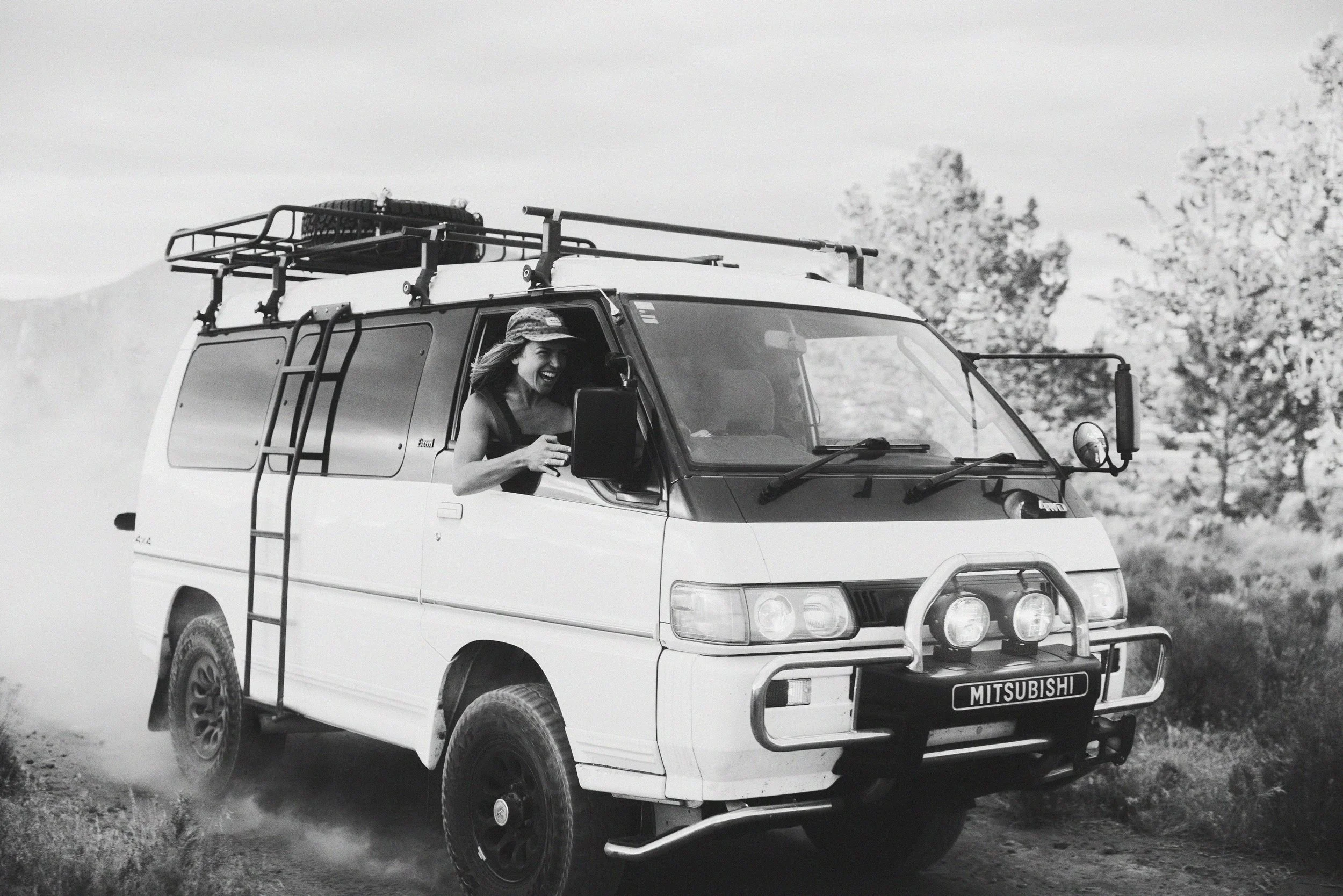 A woman with a hat smiling while driving a white Mitsubishi van on an off-road dirt path, with trees in the background and dust kicking up behind the vehicle.