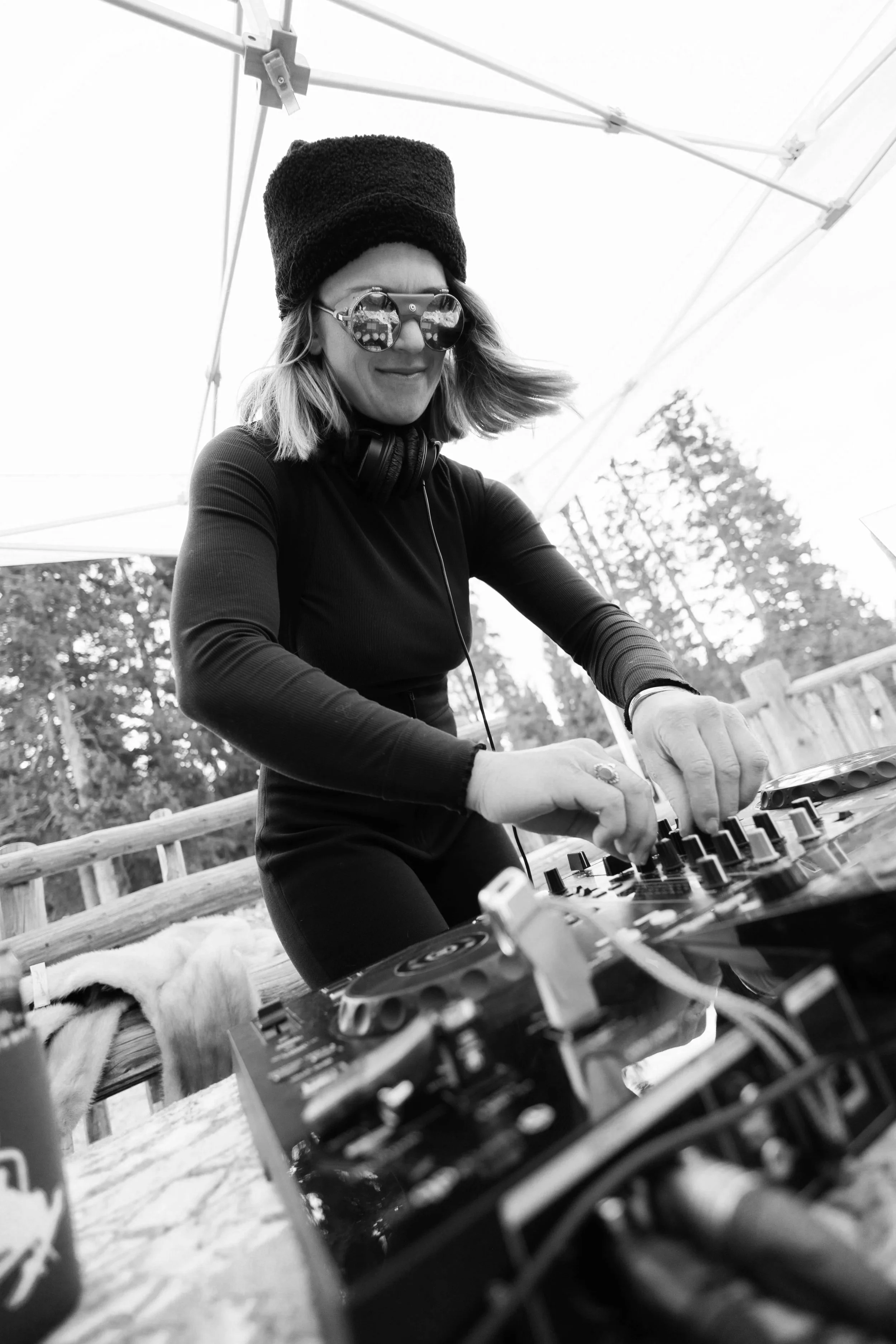 A woman wearing sunglasses, a hat, and headphones is adjusting music controls on a DJ mixer outdoors under a canopy, with trees in the background.