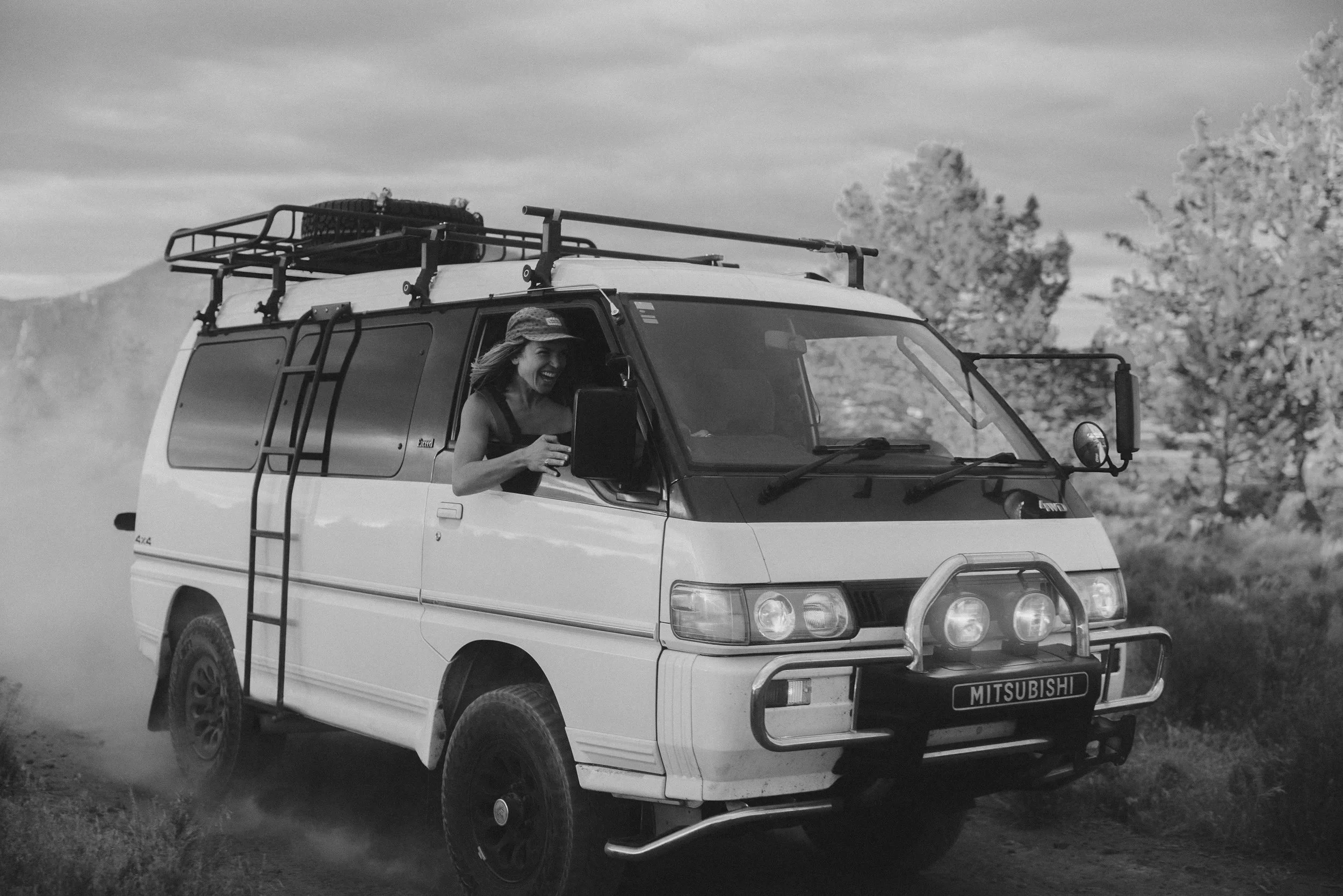 A person with a hat smiling and holding a tablet inside a Mitsubishi off-road vehicle, which is on a dirt path with trees and hills in the background, in black and white.