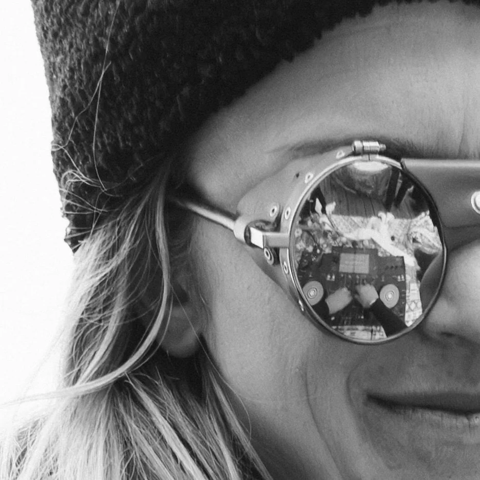 A person wearing reflective sunglasses and a beanie, with long hair, smiling and taking a selfie with a DJ mixer reflected in the sunglasses.