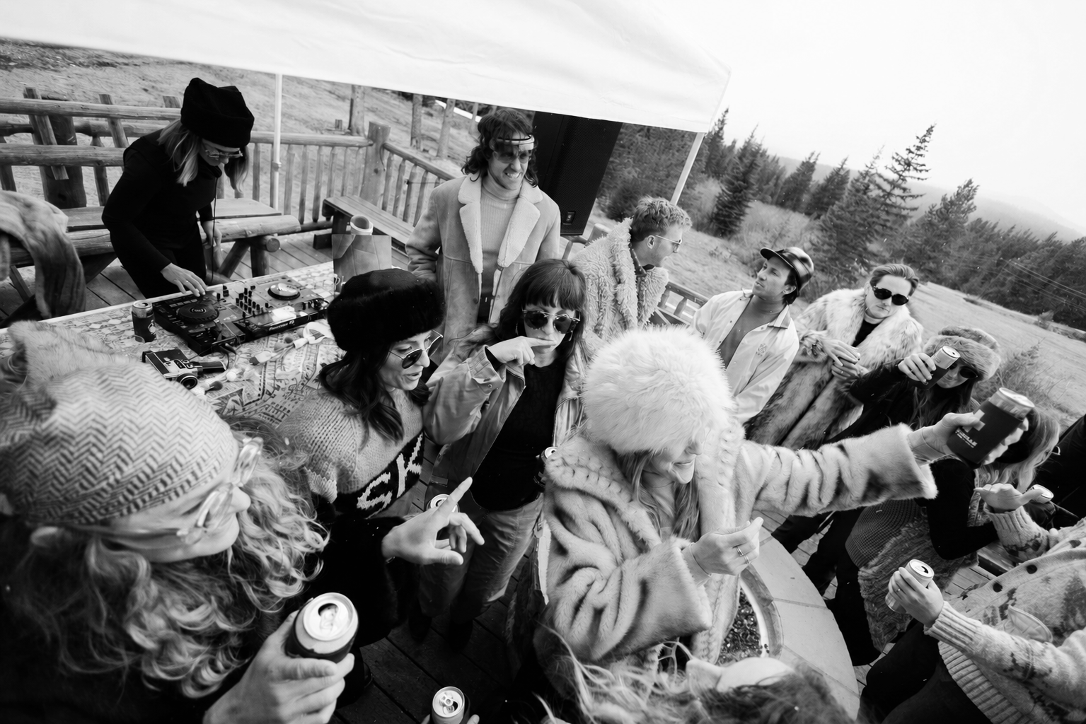 Group of people gathered outdoors under a tent, some wearing fur coats and winter accessories, enjoying drinks, taking selfies, and socializing with a DJ setup nearby.
