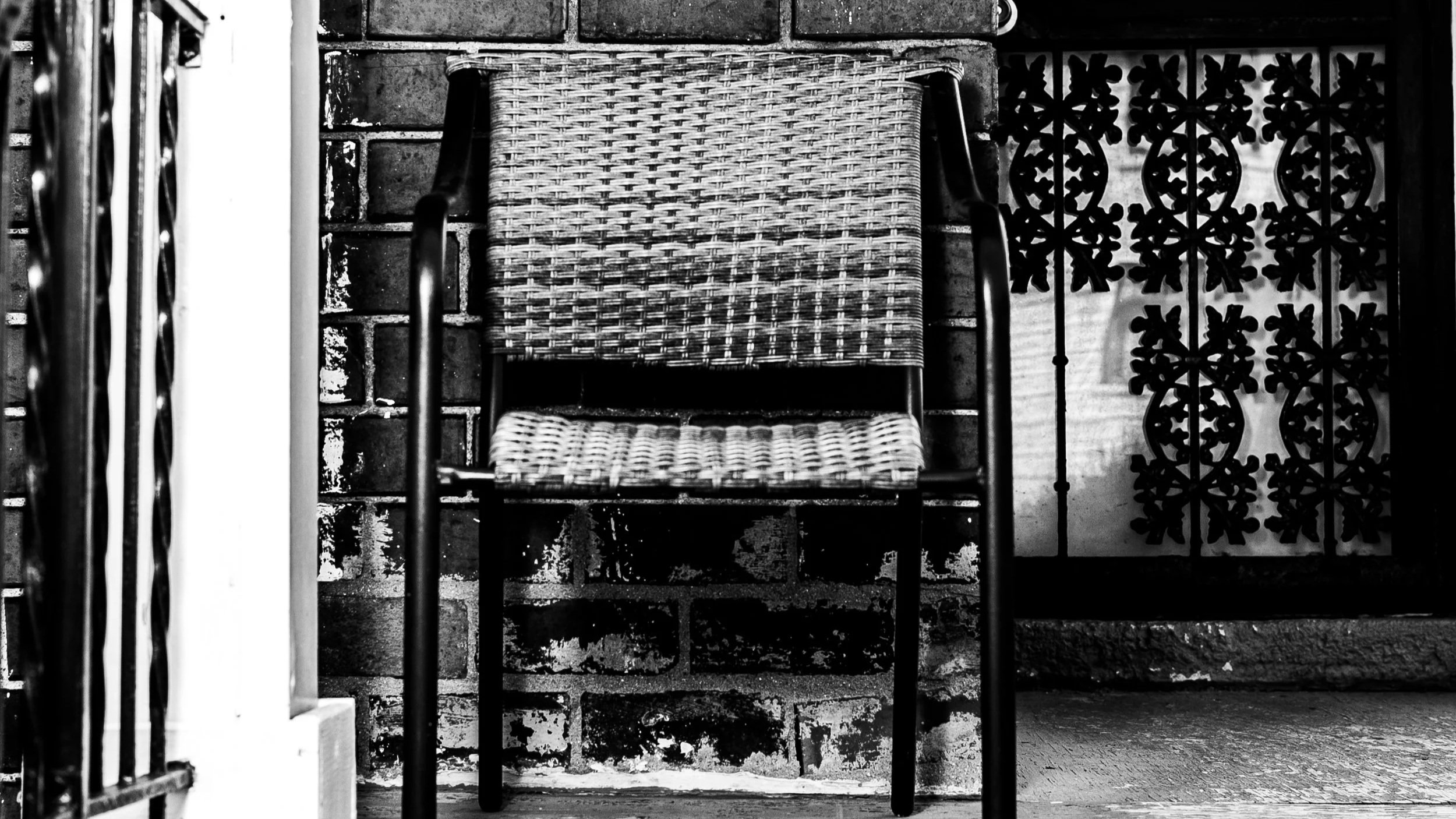 A woven wicker chair with black metal arms is positioned against a brick wall, next to decorative black wrought iron fencing with floral patterns.