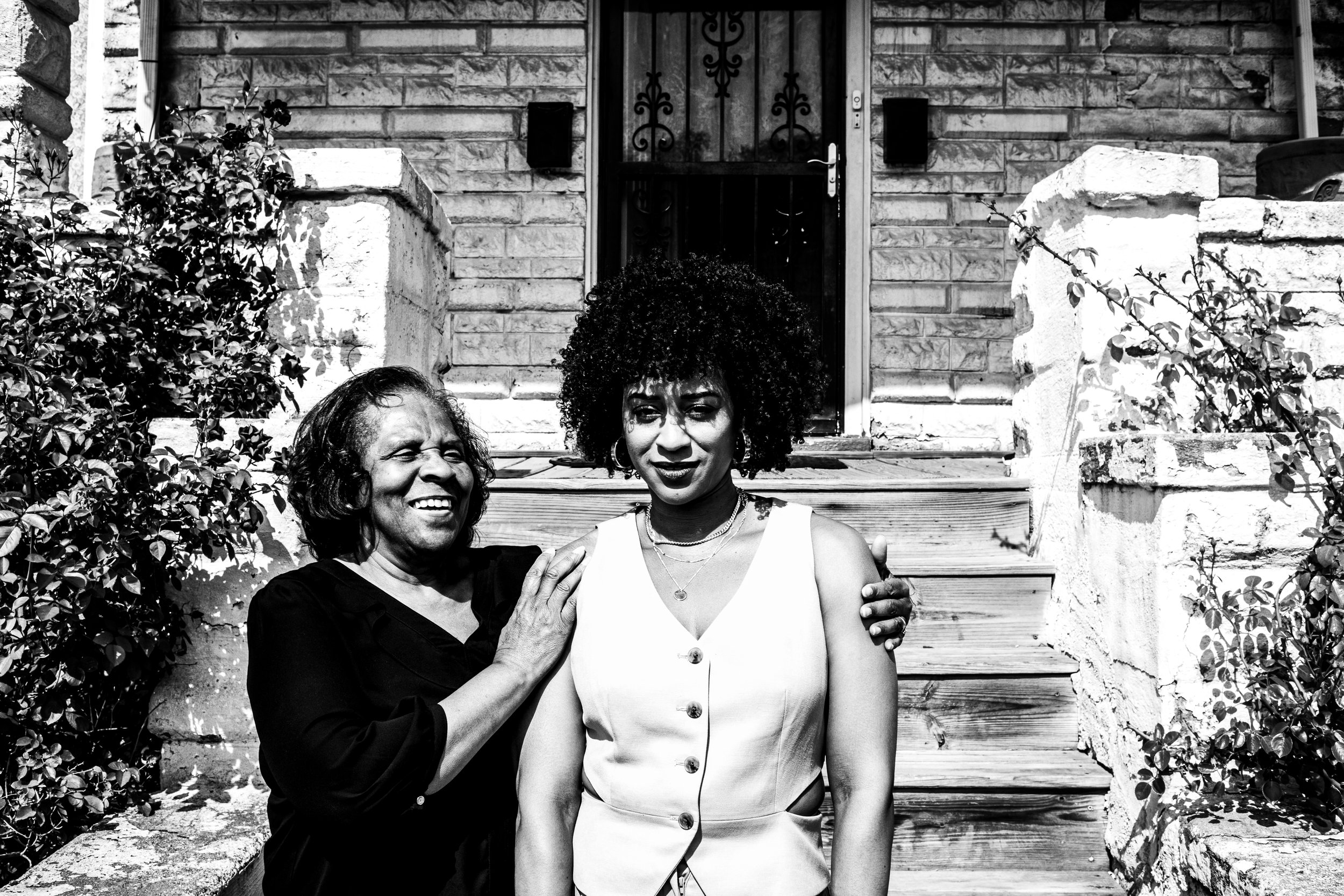 Two women standing on front porch in front of brick house, one older woman with dark short hair smiling, the other younger woman with afro hair and serious expression, both with arms around each other.