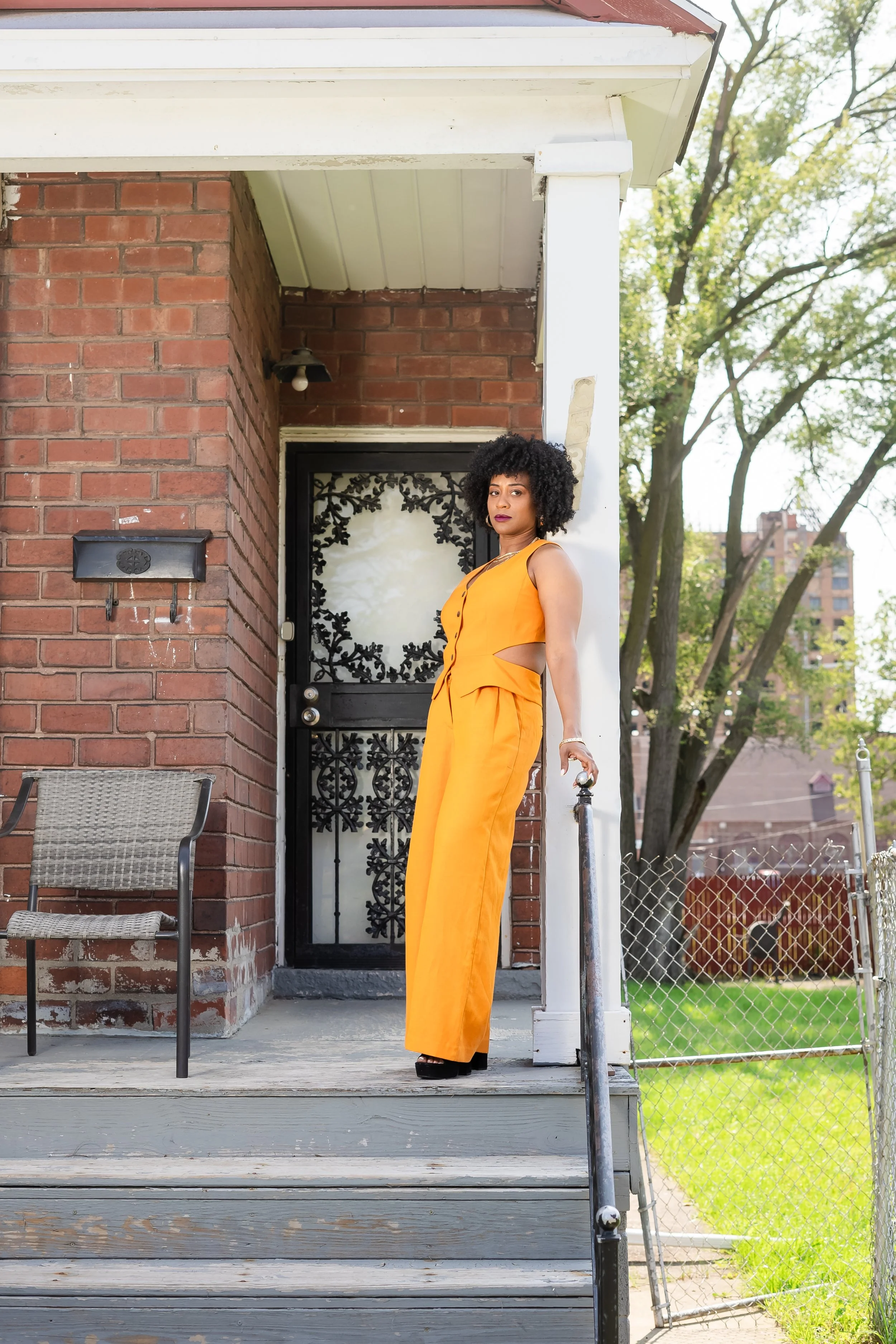 A woman with dark curly hair wearing an orange sleeveless jumpsuit standing on the porch of a brick house with a black door featuring decorative wrought iron design, a mailbox, outdoor chair, and a backyard with green grass and trees in the background.