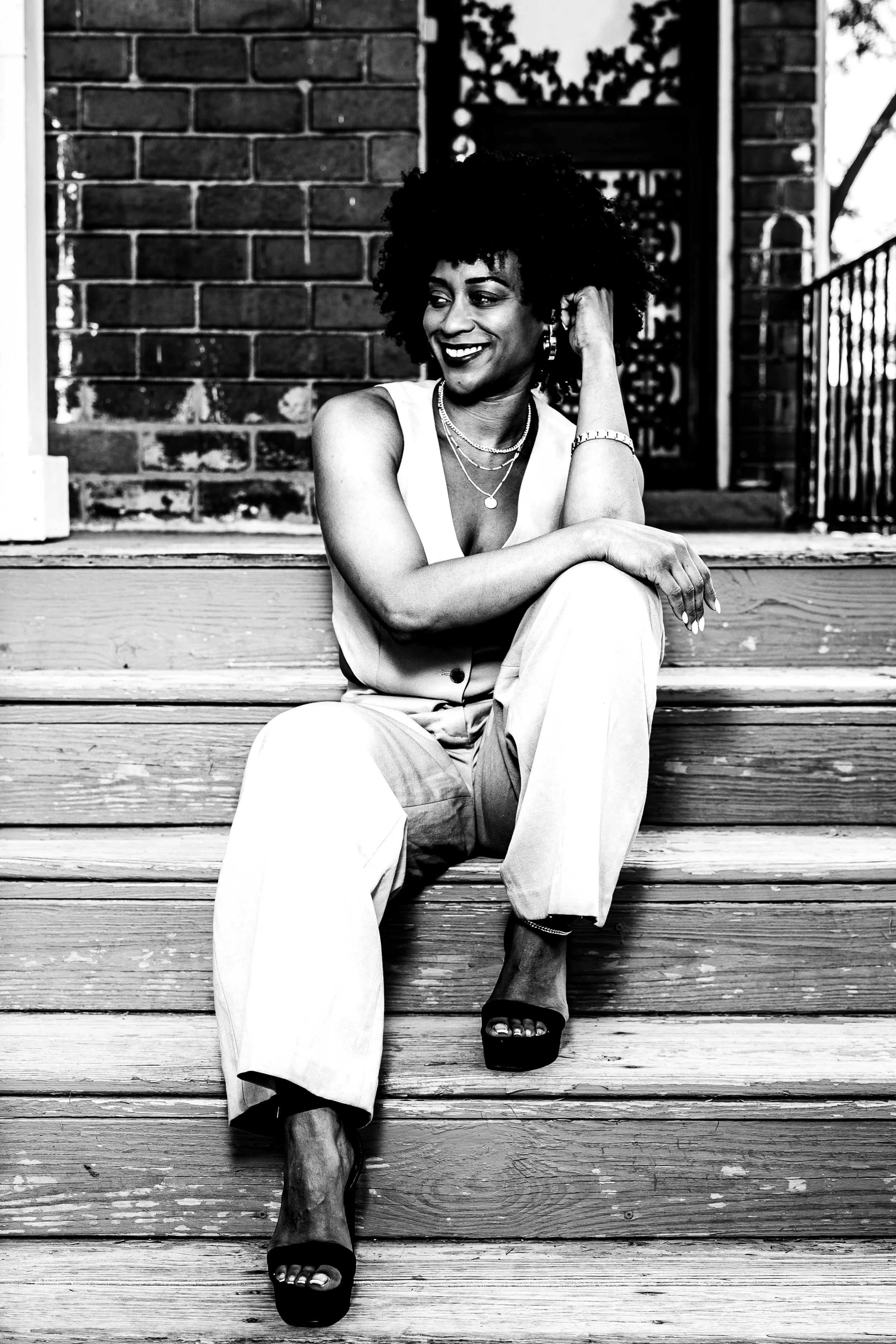 A woman sitting on wooden stairs is smiling and adjusting her earring. She has curly hair, wearing jewelry, and a sleeveless top. The background includes a brick wall and a decorative metal gate.