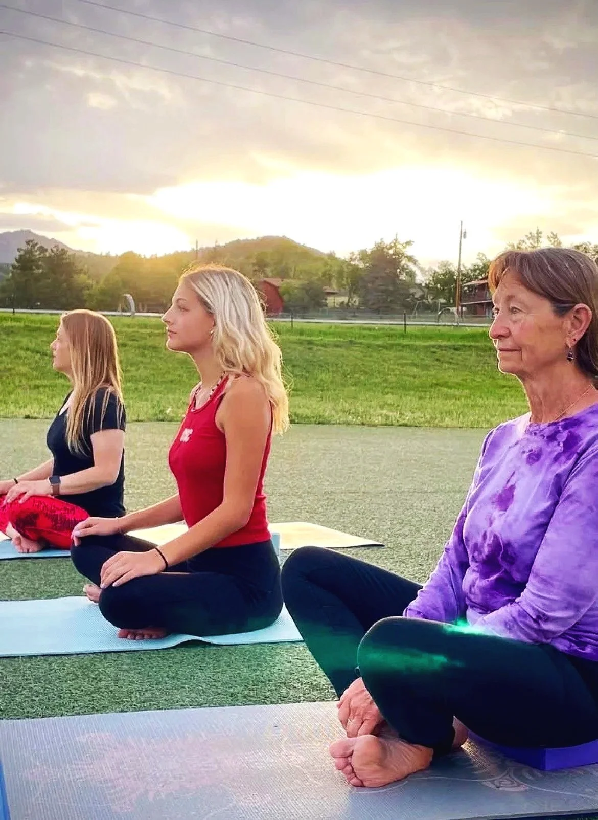 Three women and one girl practicing yoga outdoors during sunset on yoga mats, sitting in a meditative pose with legs crossed on a grassy field, with a backdrop of trees, mountains, and cloudy sky.