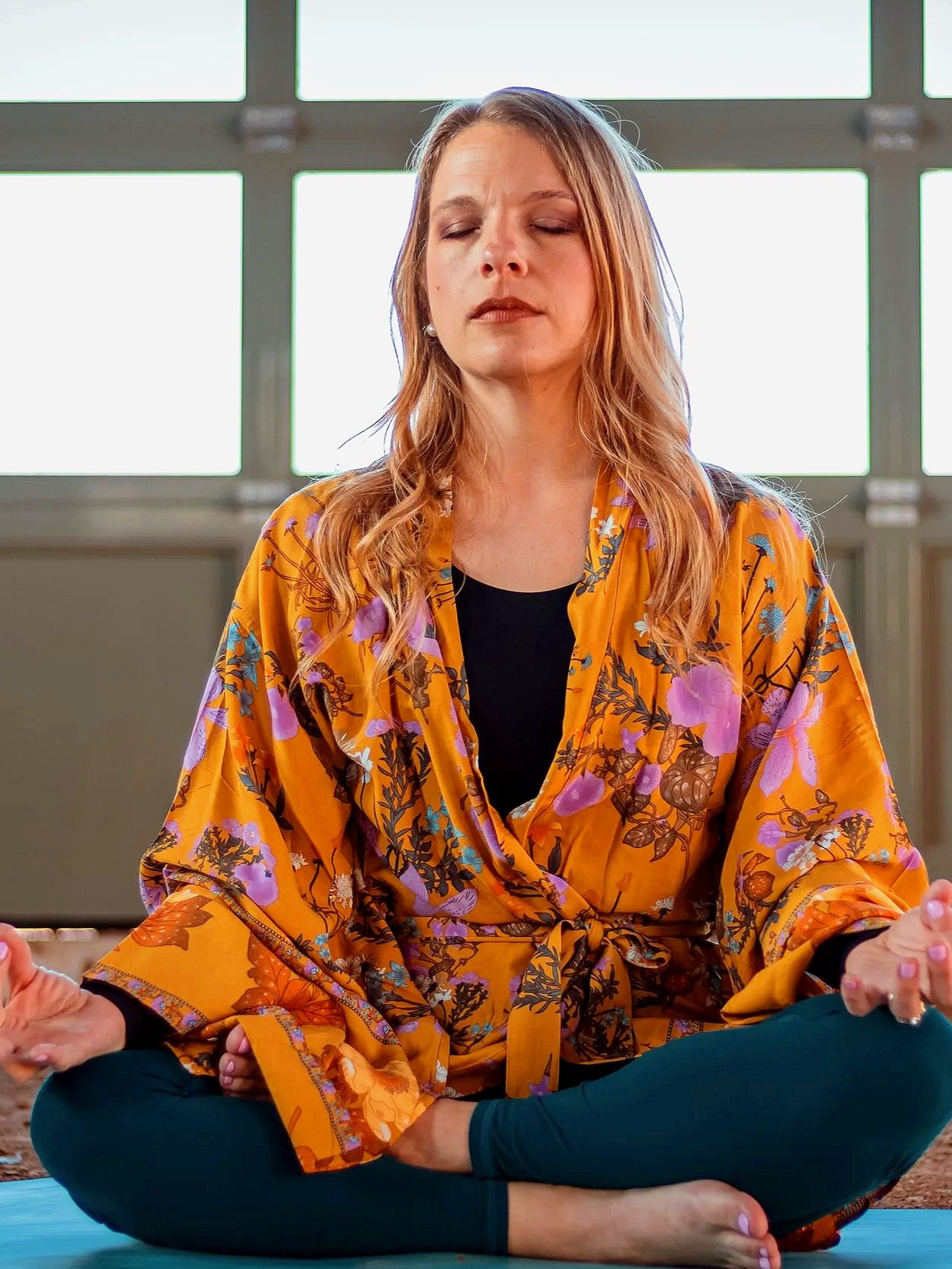 A woman practicing meditation or yoga in a sitting pose with her eyes closed, wearing an orange floral kimono, seated in front of a large window.