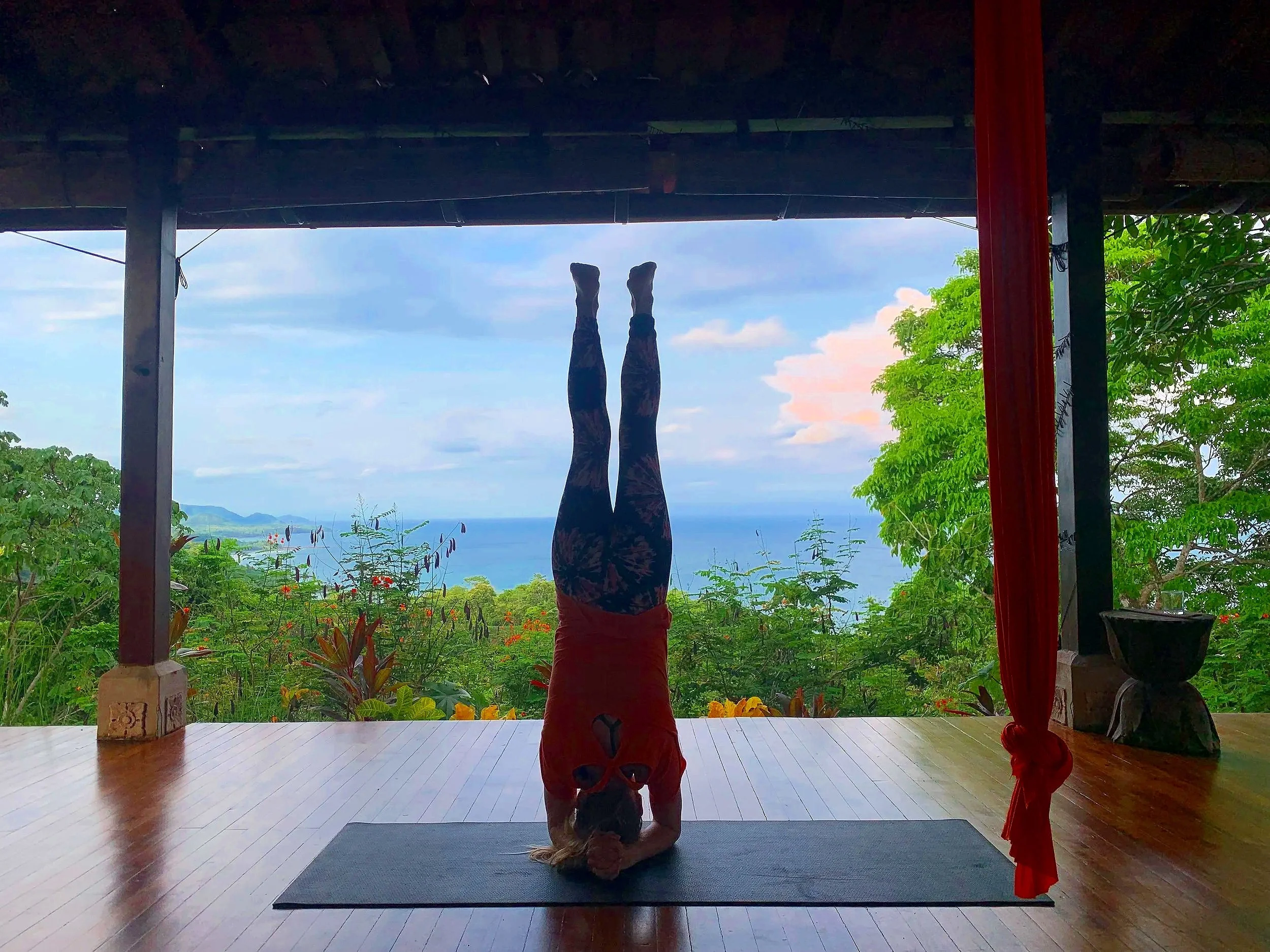 A person doing a headstand yoga pose on a black mat indoors, facing large open windows with a view of green trees, plants, and the sea in the distance.