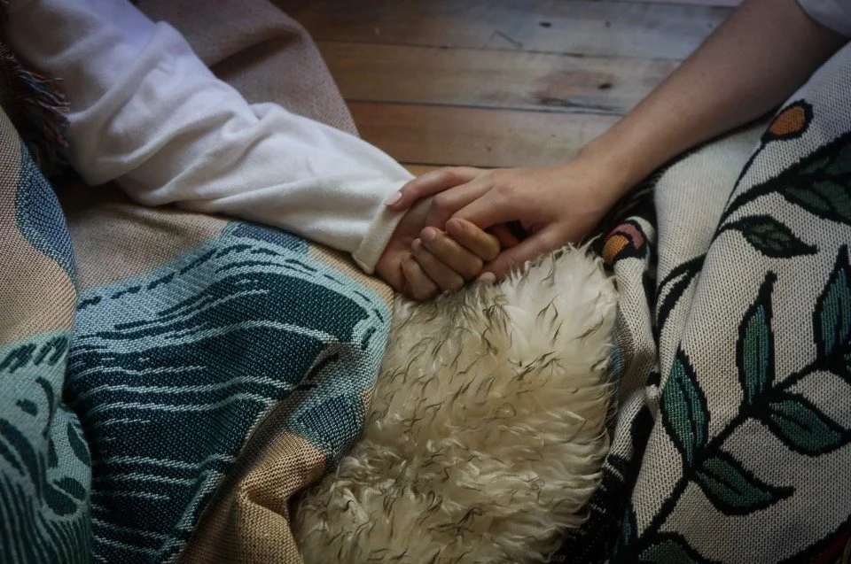 Two people holding hands, one with a light skin tone and the other with a darker skin tone, sitting on a couch with a furry blanket and patterned cushions.