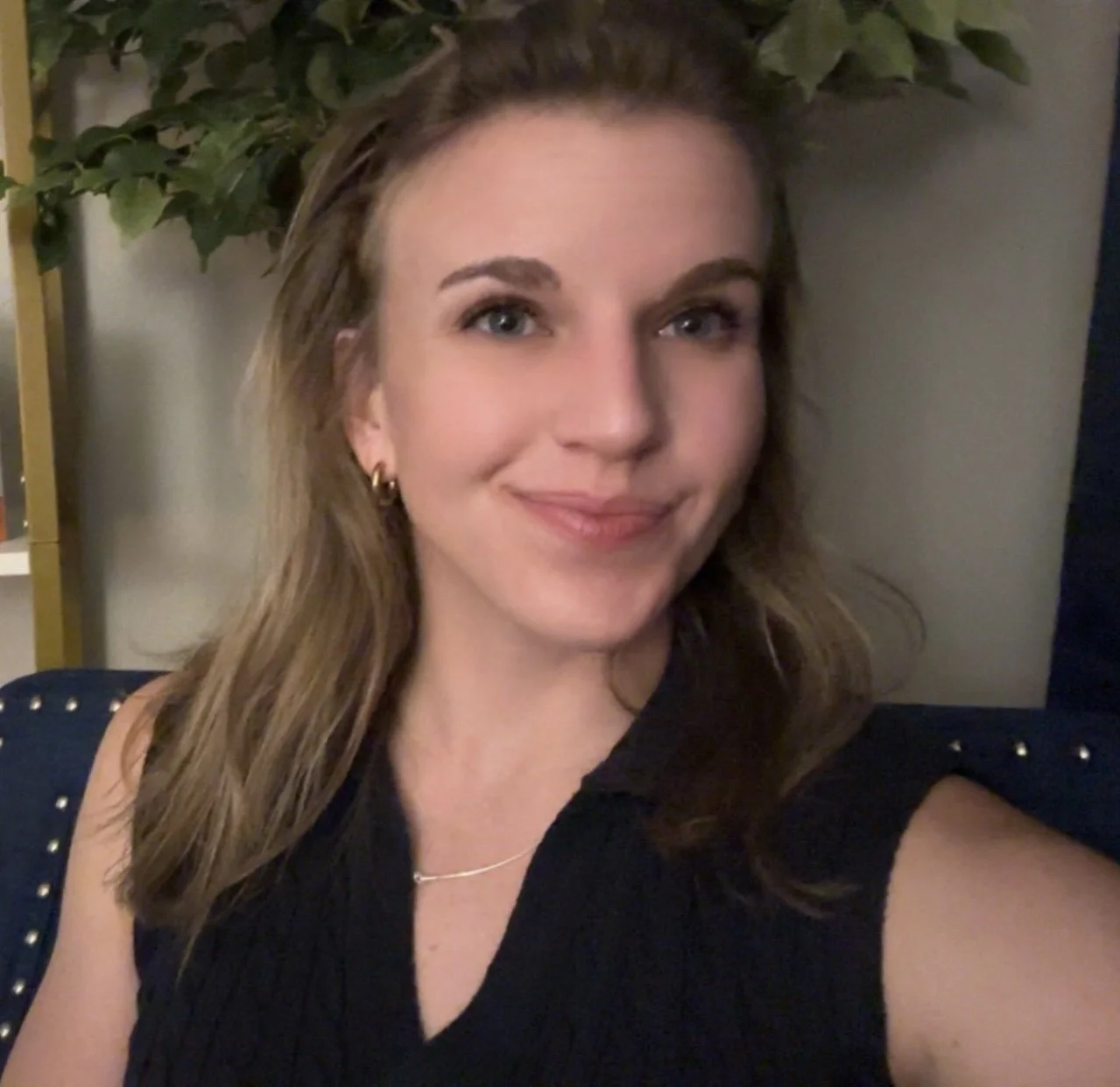 A young woman - Young therapist - with light brown hair, blue eyes, and fair skin, smiling while taking a selfie indoors. She is wearing a black sleeveless top, gold earrings, and a delicate necklace. There is greenery in the background.