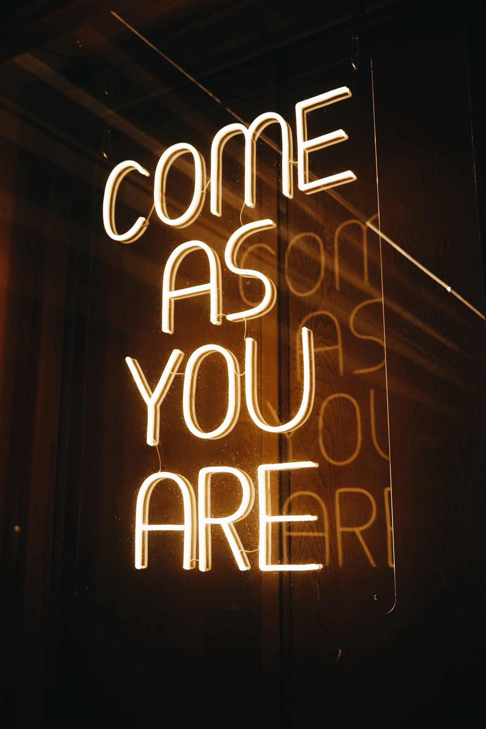 Neon sign with the words "Come Ask You Are" reflected on a glass surface.