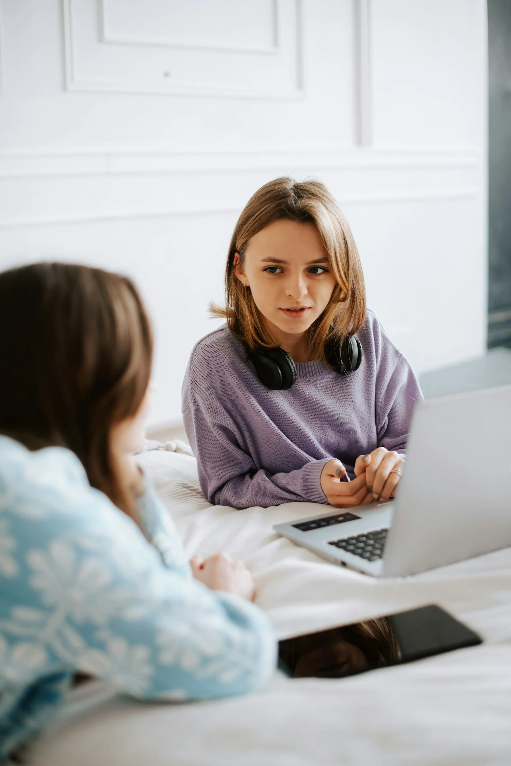 Two young women sitting on a bed having a conversation. One woman has shoulder-length light brown hair, is wearing a purple sweater, and has a pair of headphones around her neck. The other woman, seen from behind, has long dark hair and is wearing a light-colored top. There is a laptop on the bed in front of the woman with headphones.