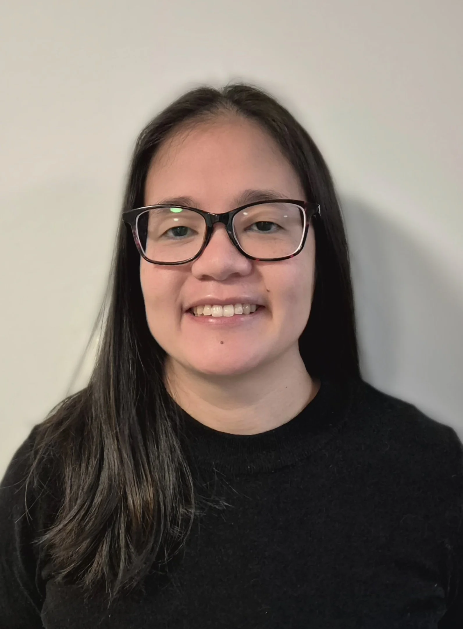 Close-up of a young woman with long dark hair, wearing glasses and a black top, smiling at the camera against a plain white wall background.