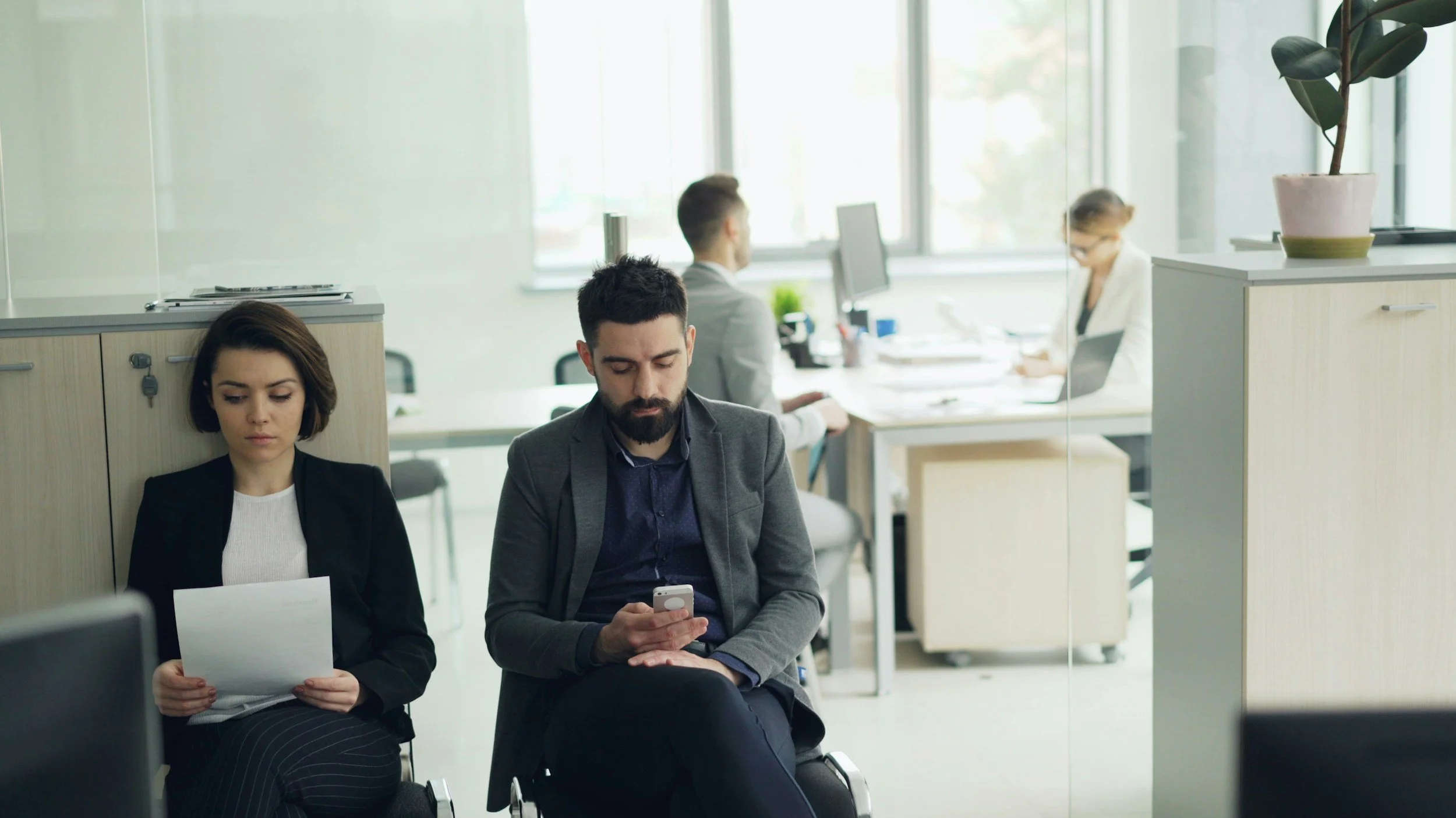 Deux personnes assises dans une salle d'attente d'entreprise, une femme avec une feuille et un homme regardant son téléphone, avec d'autres employés travaillant à des bureaux en arrière-plan.