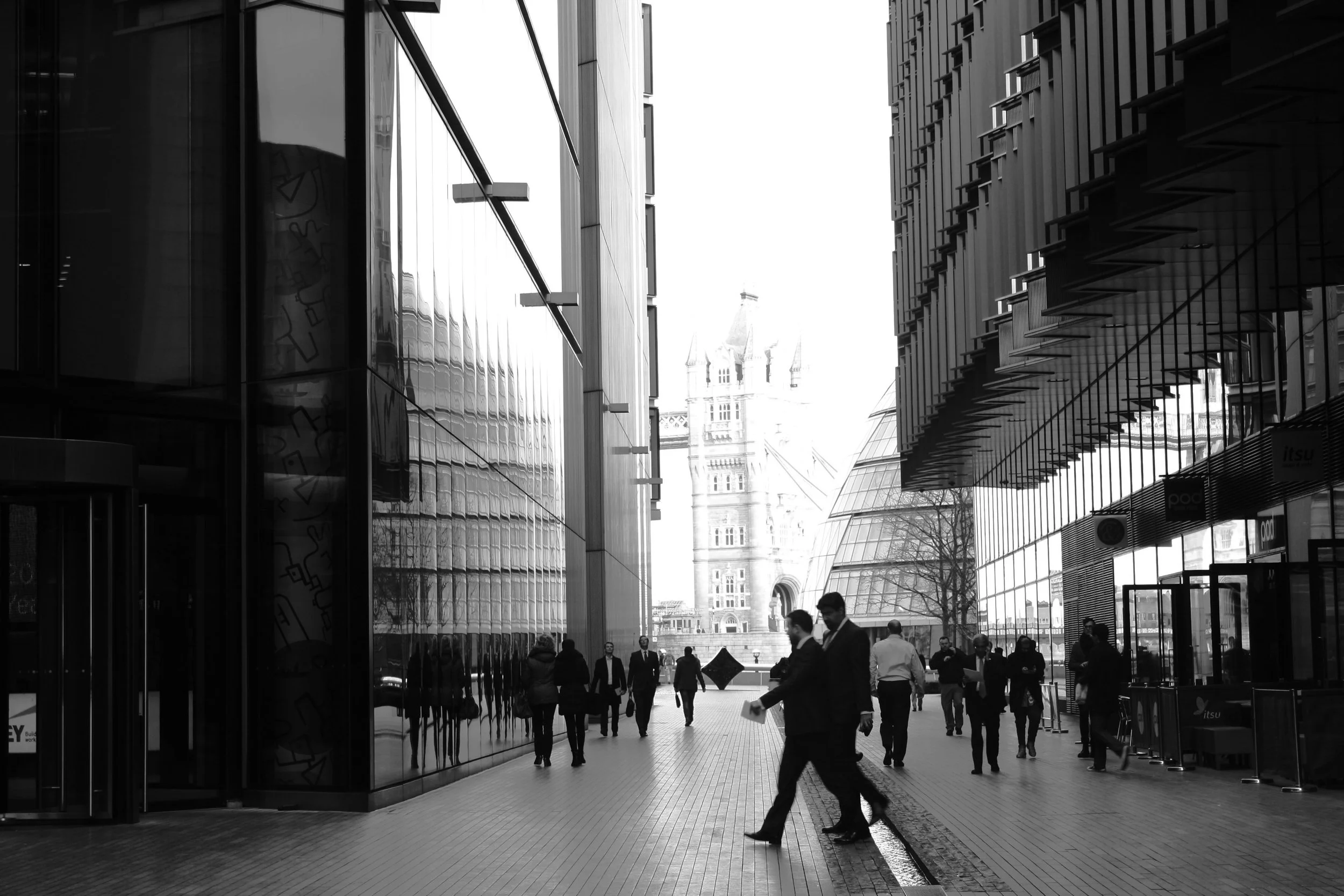 Photo en noir et blanc d'une rue urbaine avec des bâtiments modernes en verre à gauche et à droite, et un pont en arrière-plan, avec des personnes marchant dans la rue.