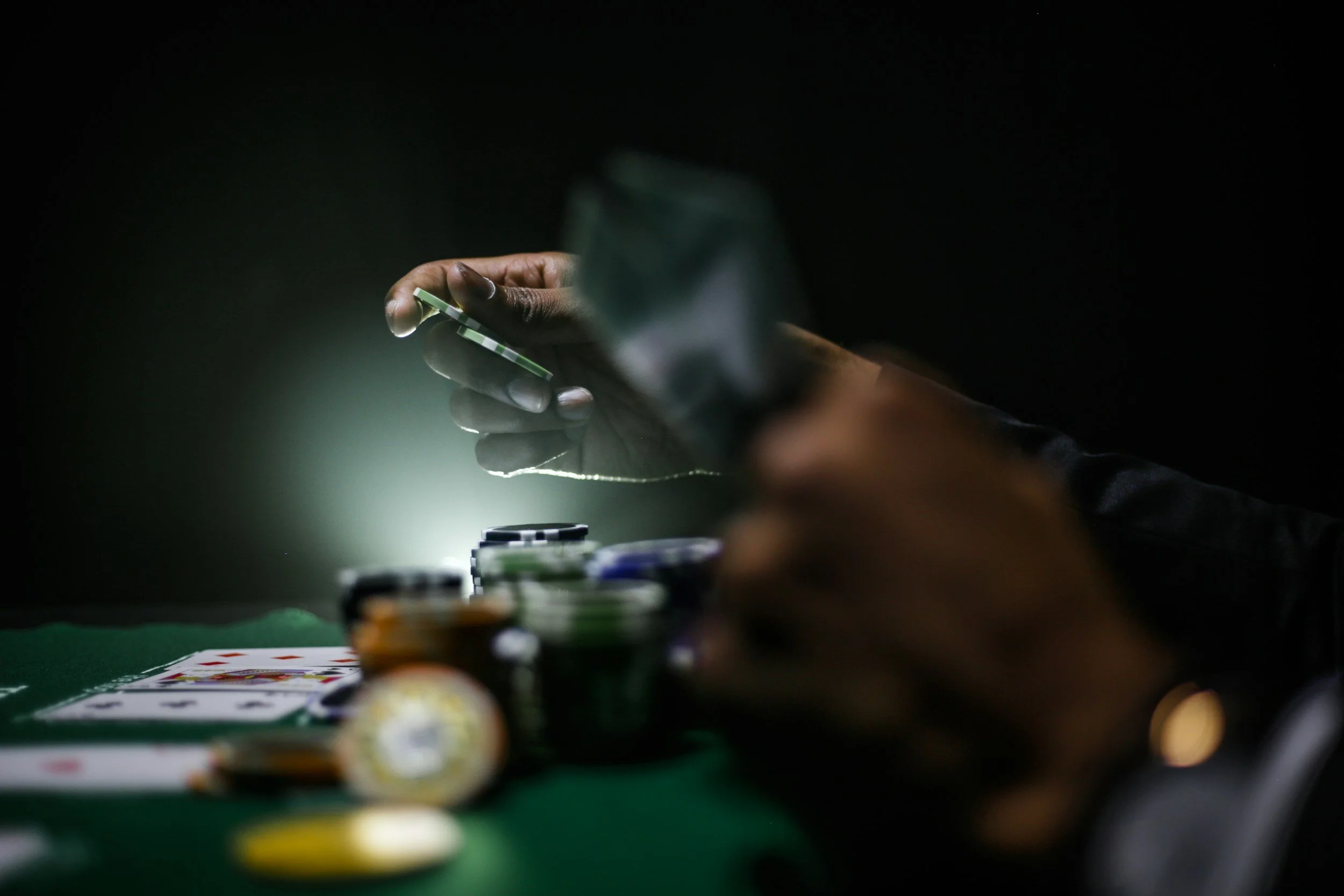 A person with dark skin holding a green poker chip over a poker table, with stacks of chips and playing cards visible in the background.