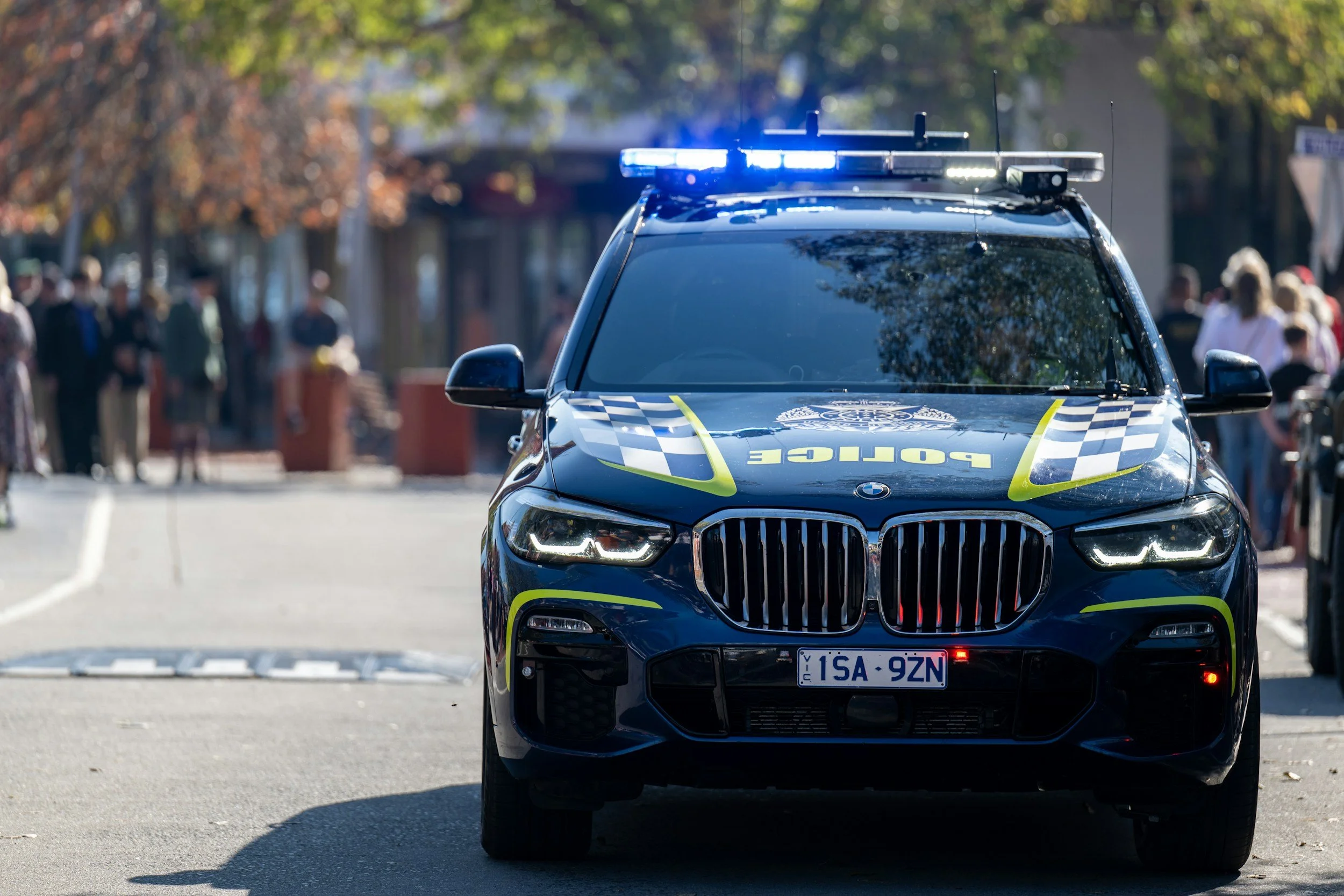 A police car with blue emergency lights on, parked on a street with people walking and trees in the background.