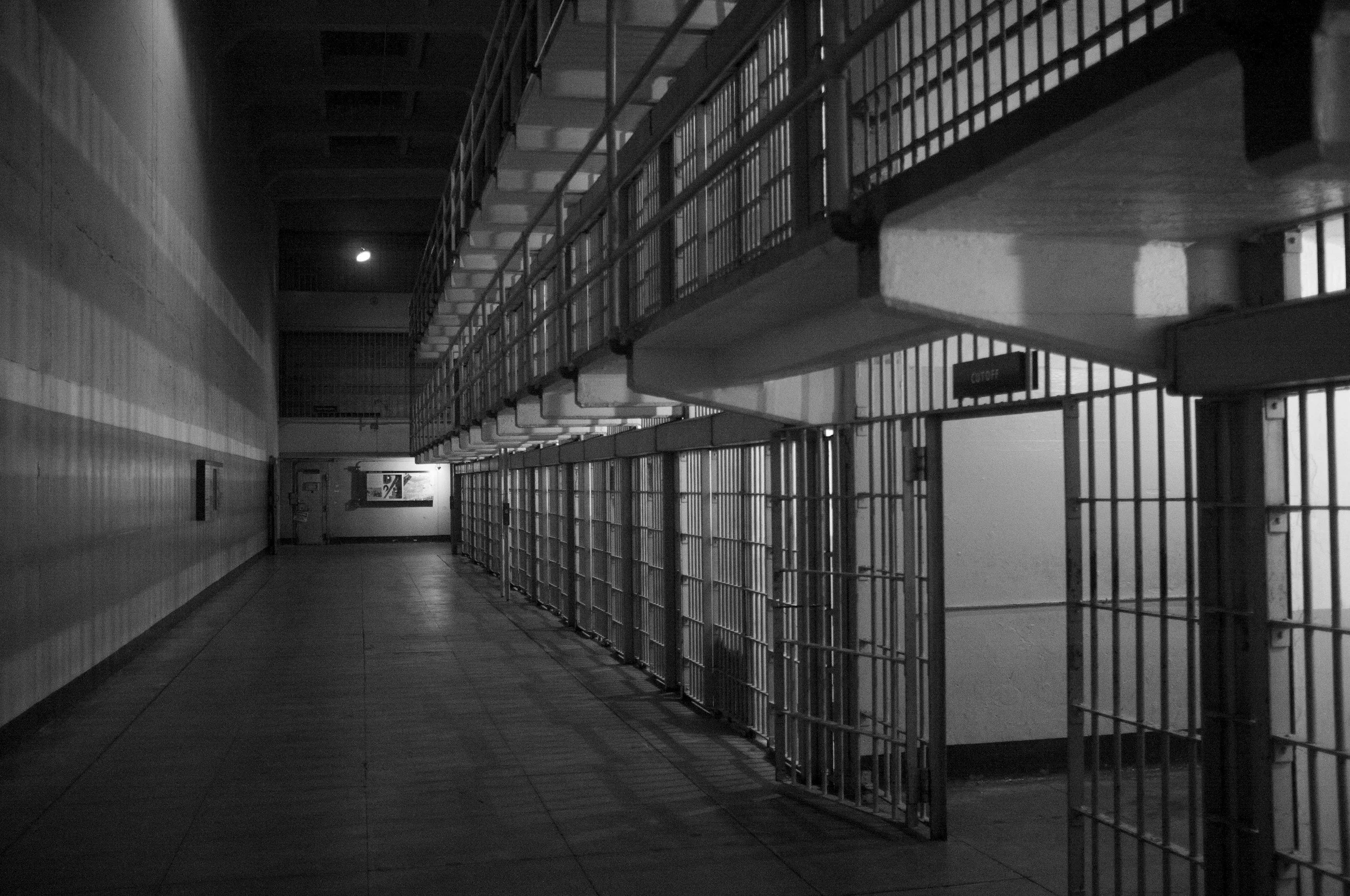 Black and white image of a prison corridor with metal cell bars and locked doors on the right side, dim lighting, and a moon visible through a window.
