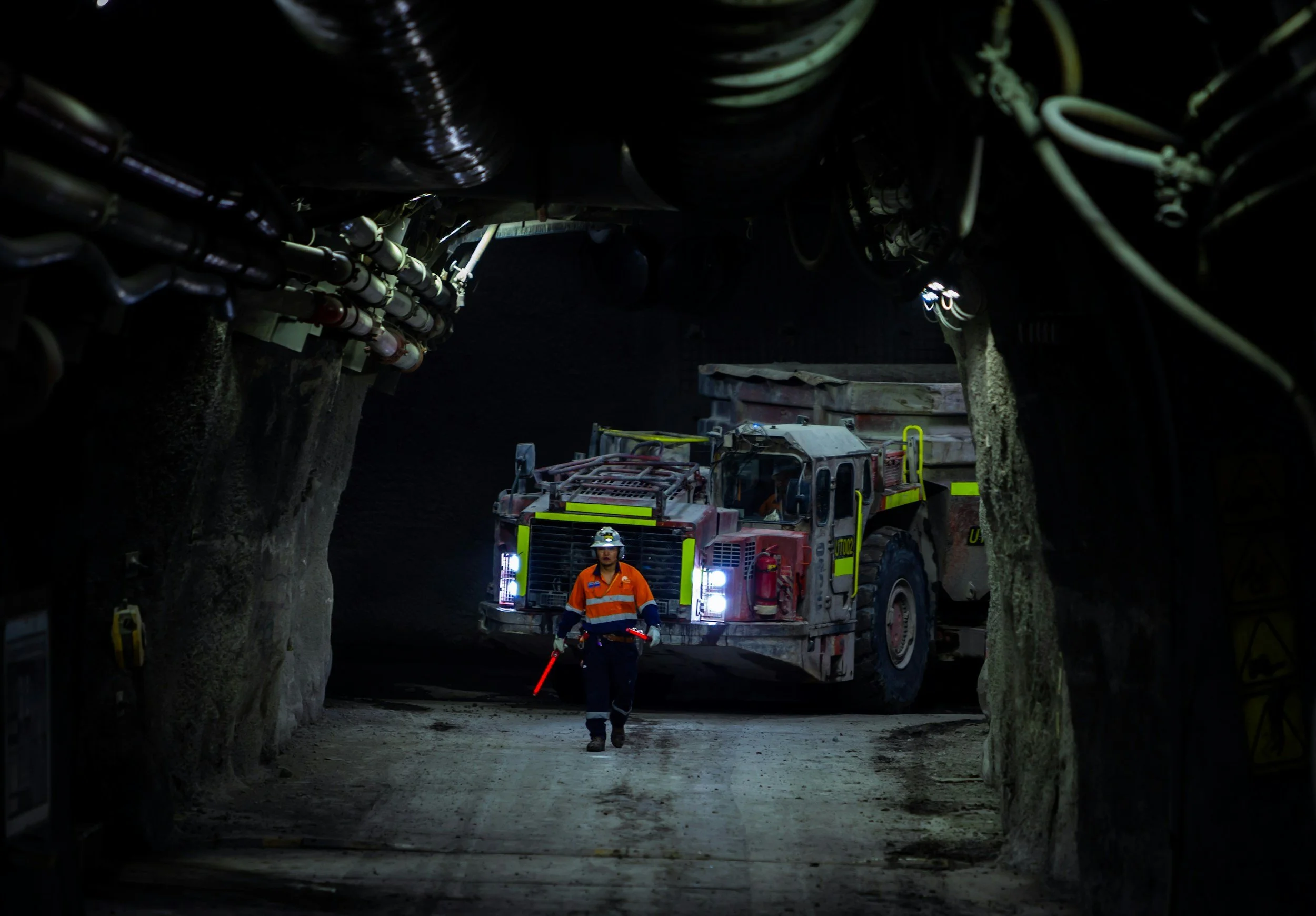 A firefighter in orange uniform walking in front of a fire truck inside a tunnel under construction, surrounded by rock walls and pipes, with a worker walking behind.
