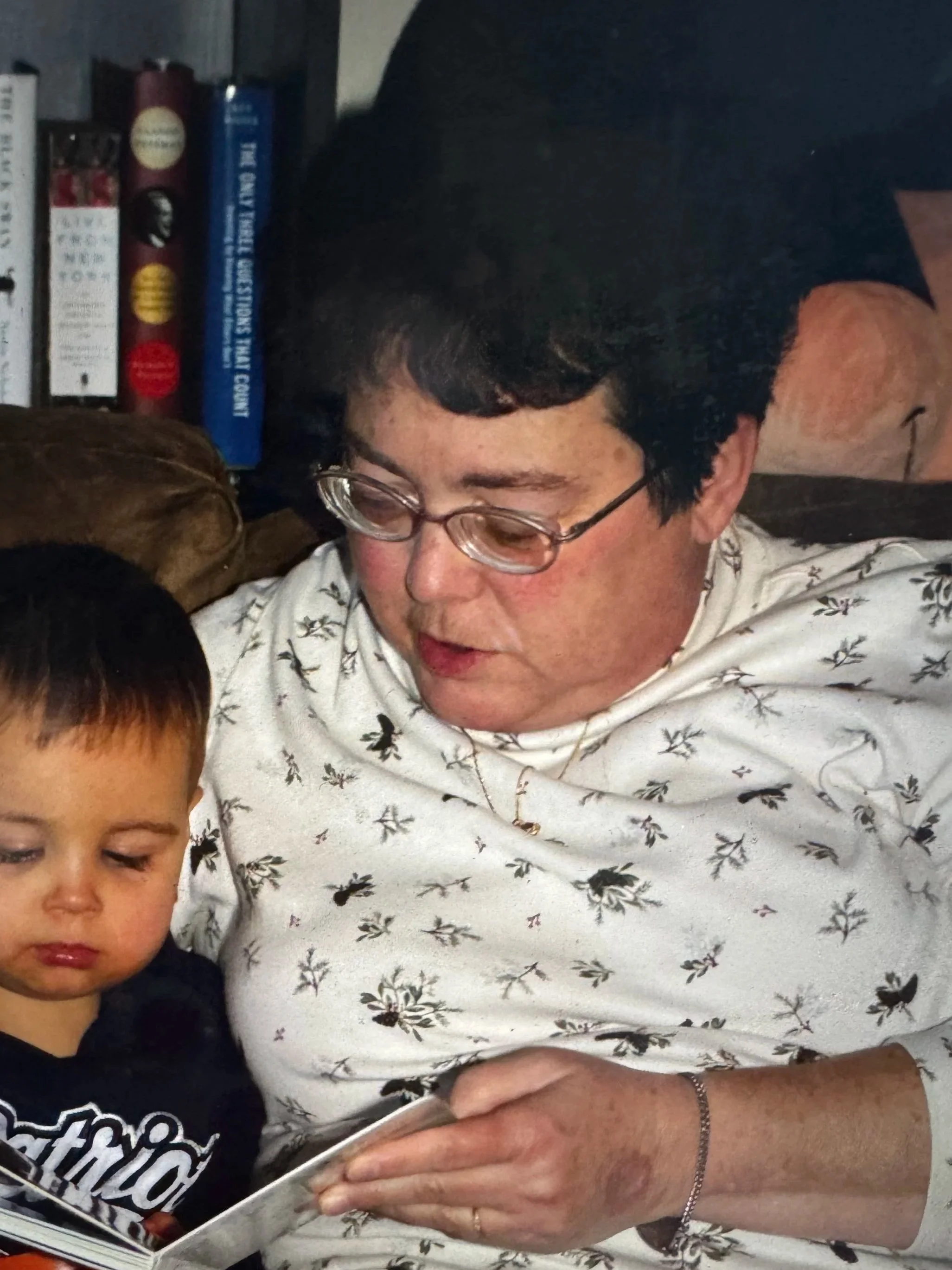 A woman with short black hair and glasses reading a book, with a young boy partially visible in the foreground, in a room with a bookshelf in the background.