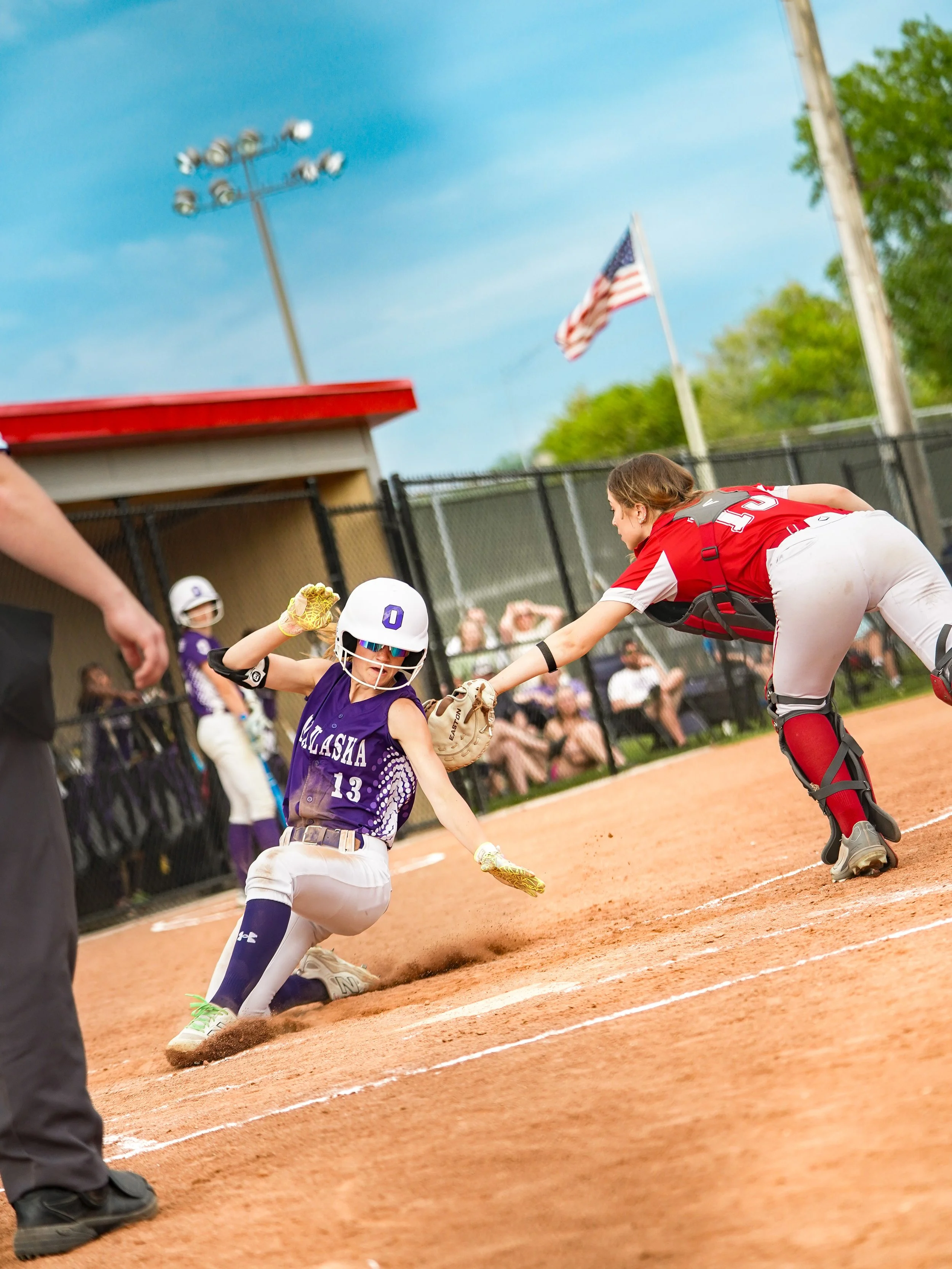 A young girl sliding into home base during a baseball game, wearing a purple jersey with the number 13, and a baseball helmet with an O on it, while a catcher in a red uniform waits to tag her out.