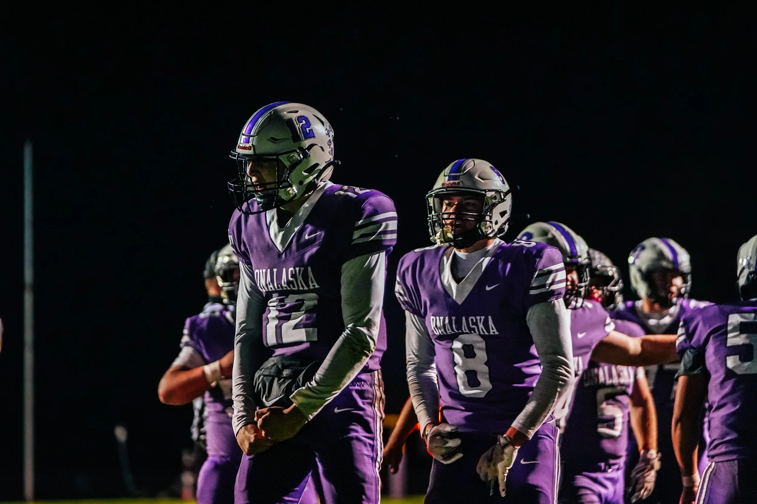 Alaska football team huddled together during a nighttime game, wearing purple jerseys and helmets.