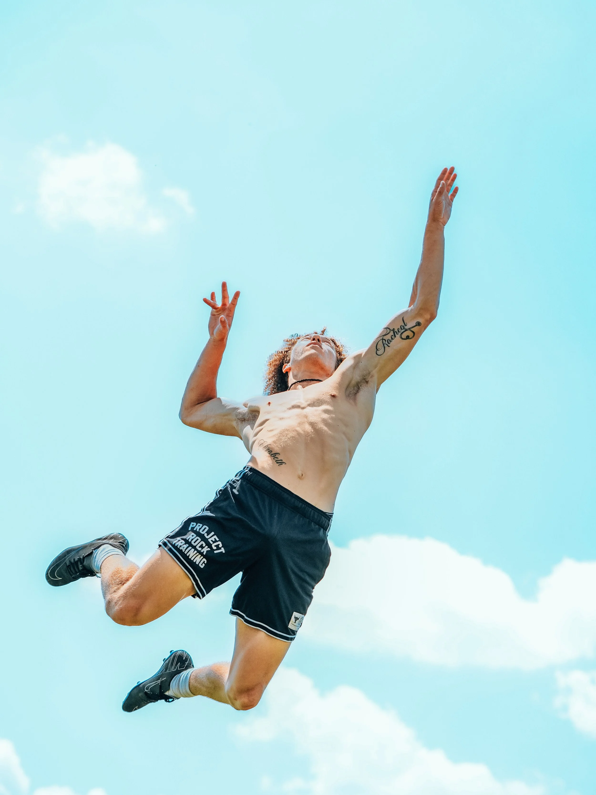 A shirtless man with tattoos jumping in the air against a blue sky with clouds, wearing black shorts and running shoes.