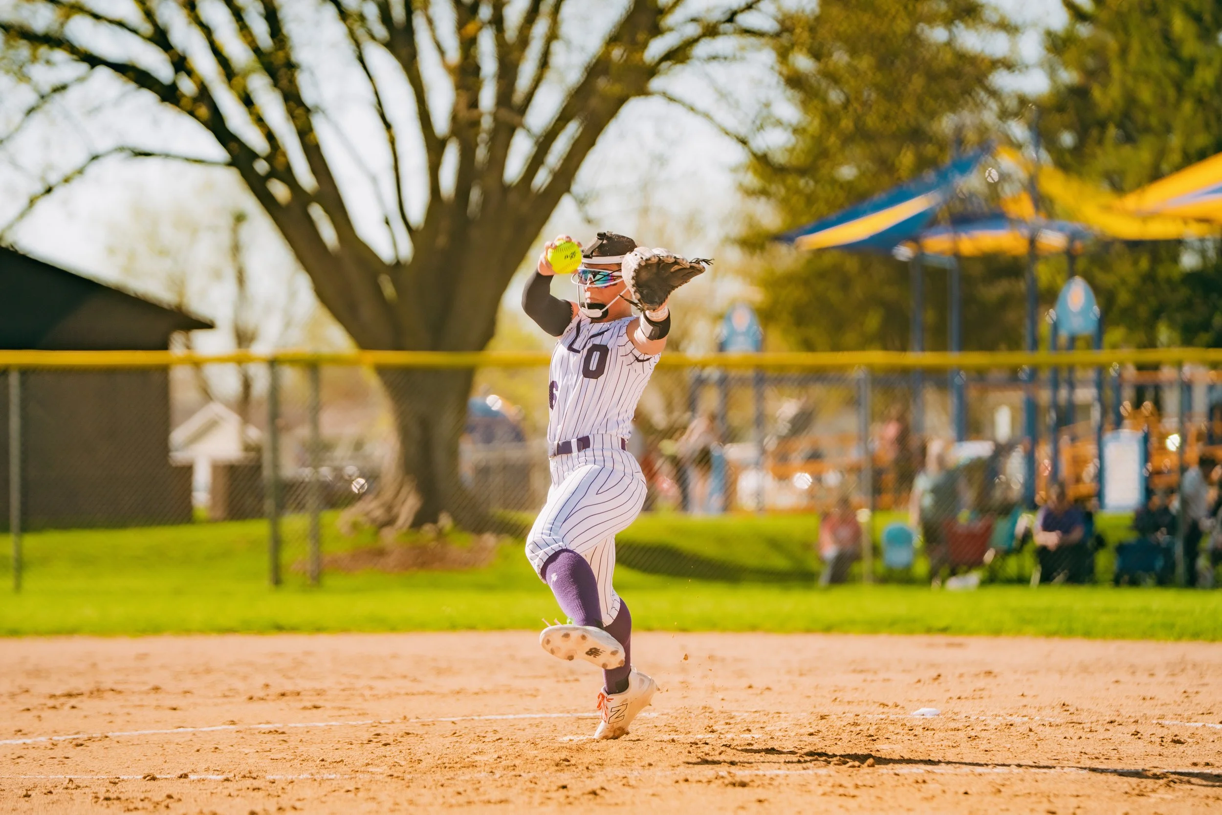 A softball player in a striped uniform is running on the field with a baseball glove, holding a yellow softball in her right hand, and a baseball bat on her shoulder. She is wearing helmet, sunglasses, and knee-high socks. There are trees and playgro