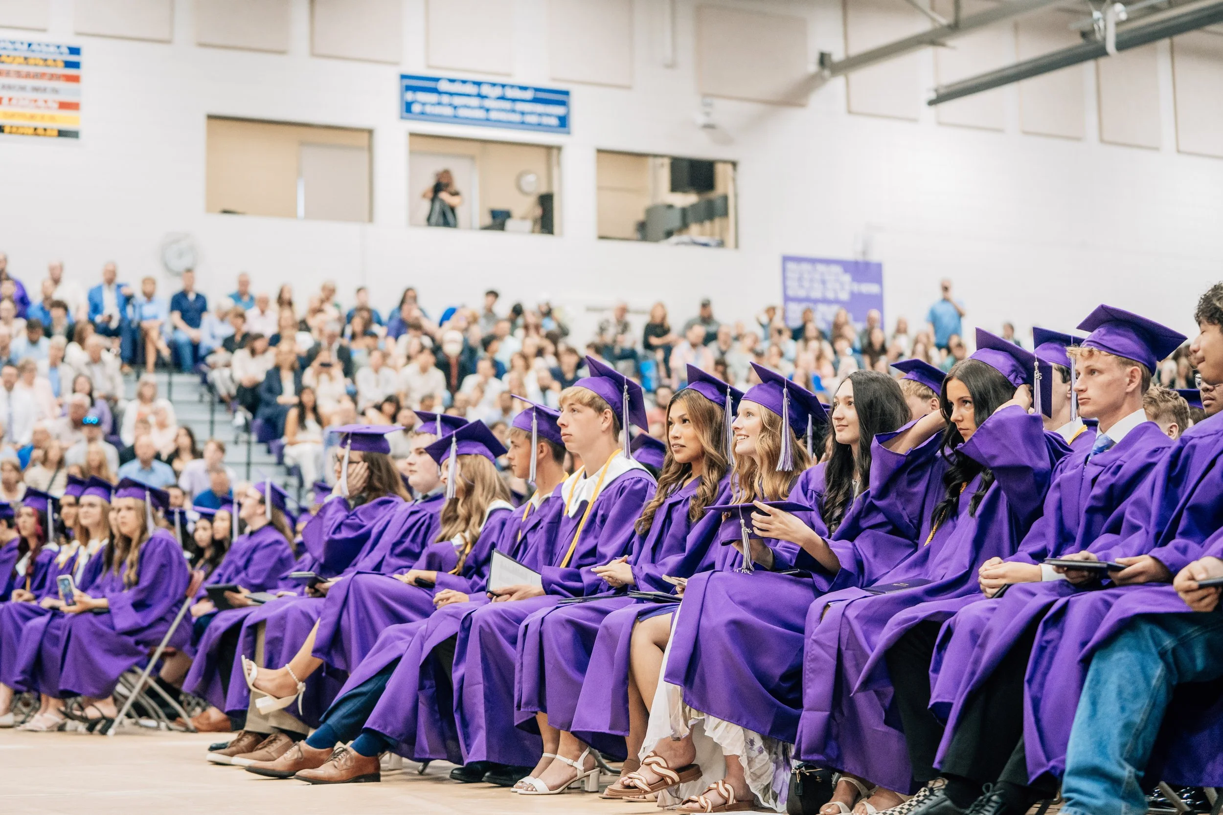 Group of graduates wearing purple caps and gowns attending a graduation ceremony in an indoor gymnasium with seated audience and blue banners.