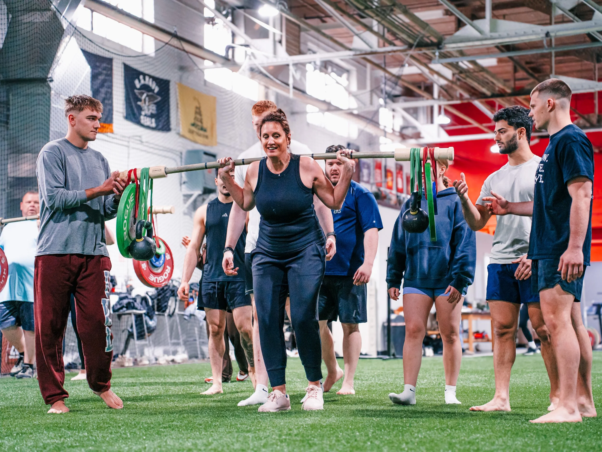 A group of people engaging in a fitness class in an indoor gym, with a woman lifting a barbell while others observe or instruct.