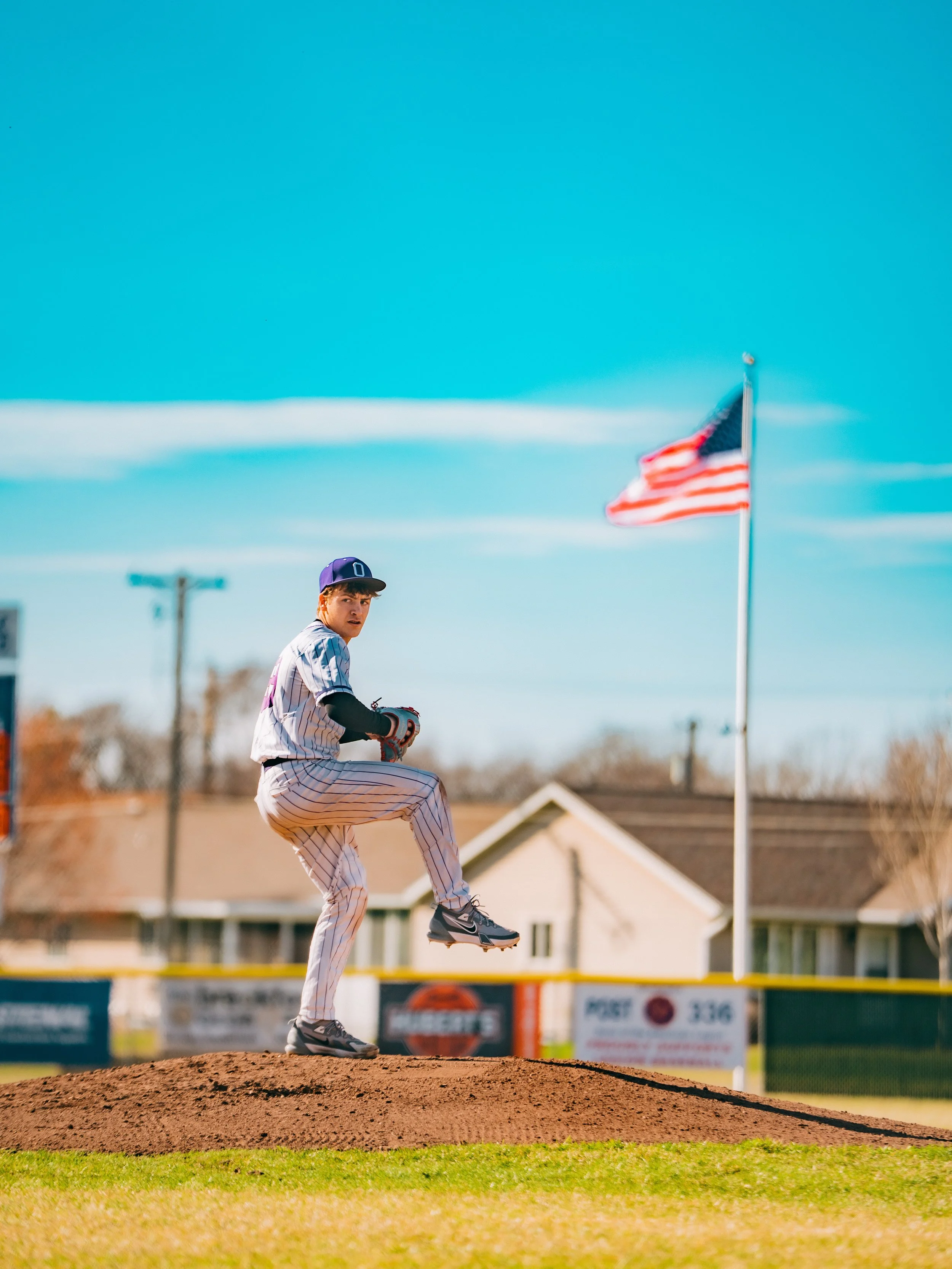 A young baseball player in a jersey and cap is pitching on the mound during a game, with a blue sky overhead and an American flag waving nearby.
