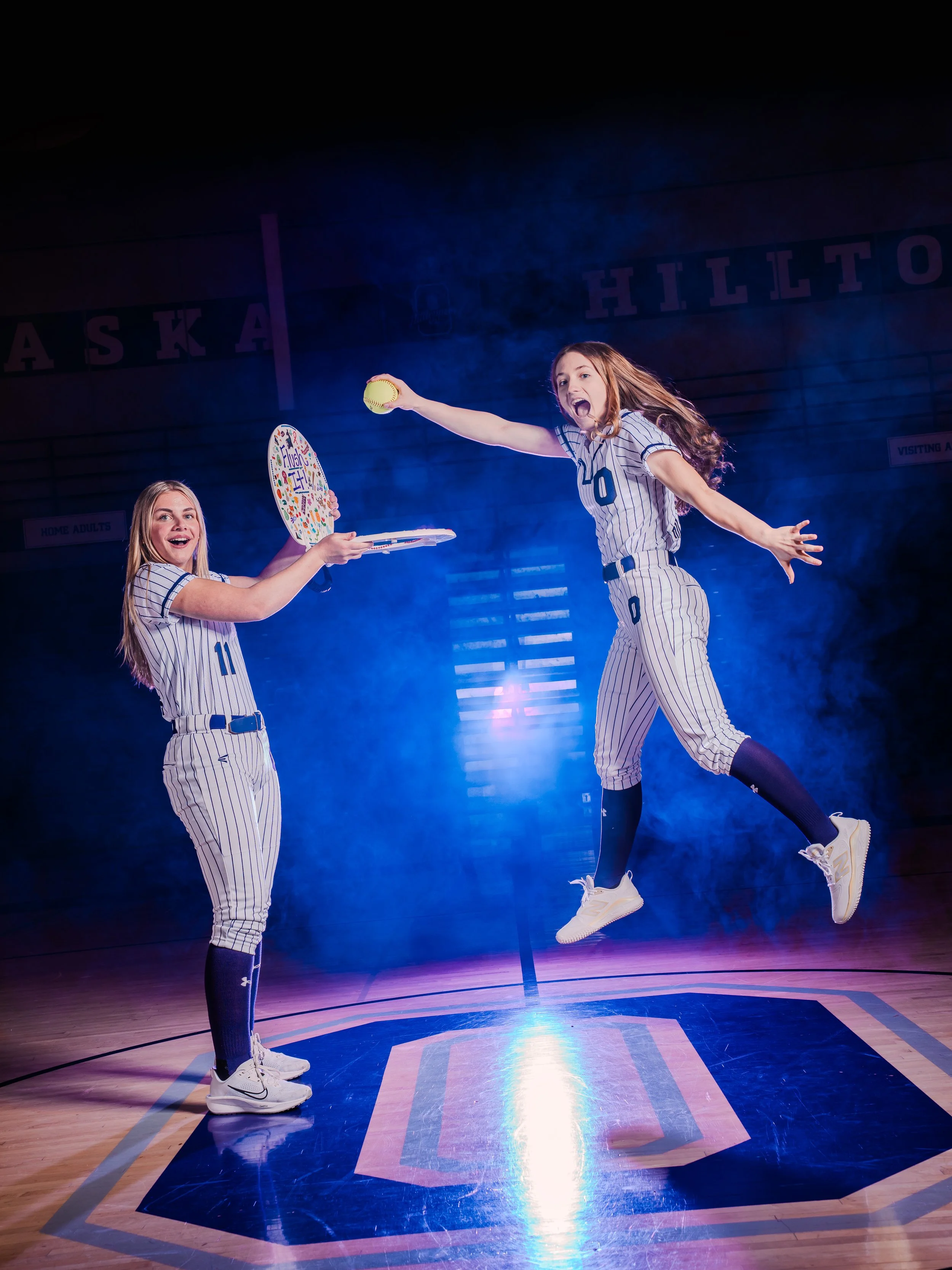 Two young girls in basketball uniforms are on a basketball court with a dark background and blue lighting. One girl is jumping while holding a yellow softball, and the other girl is smiling and holding a colorful foam tray. Both girls are wearing str