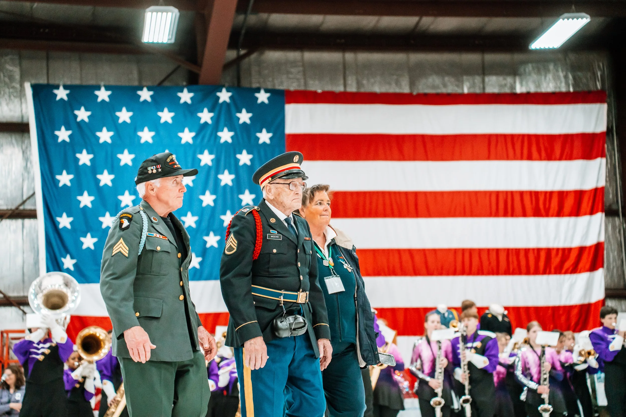 Three elderly veterans in military uniform standing with their hands at their sides, in front of an American flag backdrop, during a patriotic event. Behind them, a marching band and a children's choir are visible.