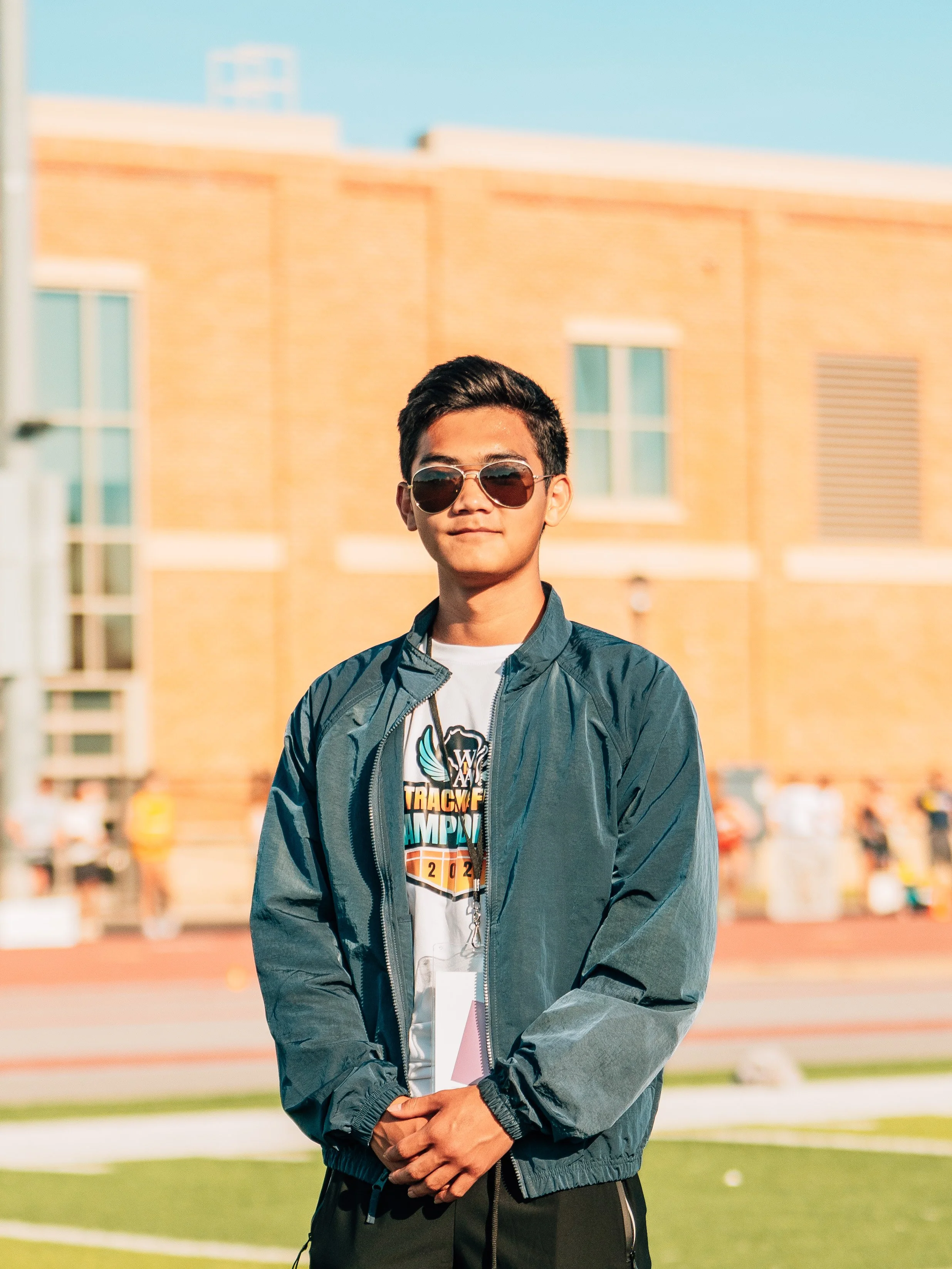 A young man with dark hair wearing sunglasses, a blue jacket, and a printed t-shirt standing outdoors on a sports field during daytime.