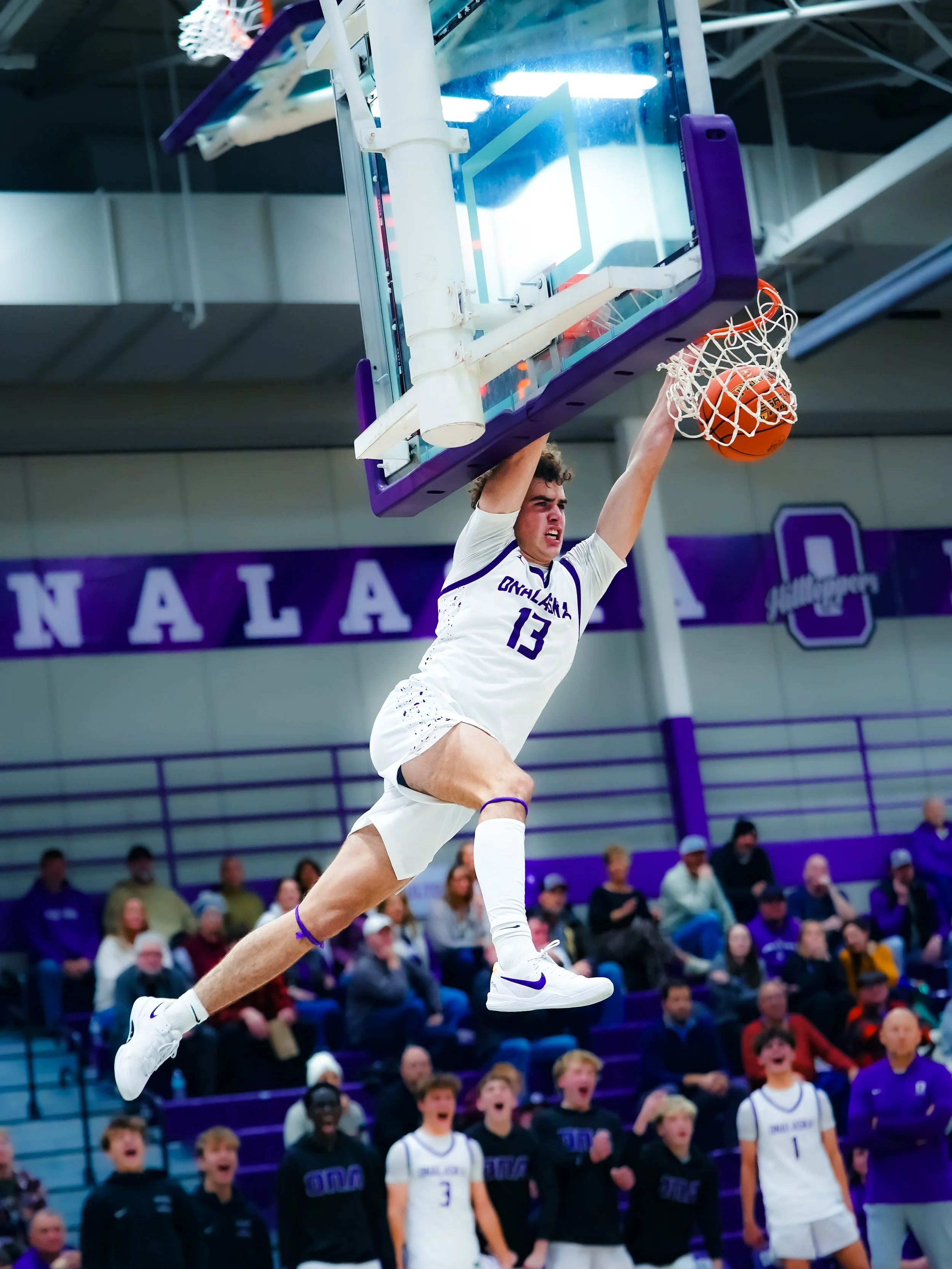 A basketball player wearing a white jersey with purple accents, numbered 13, performing a slam dunk during a game. Spectators are visible in the background, watching the action.