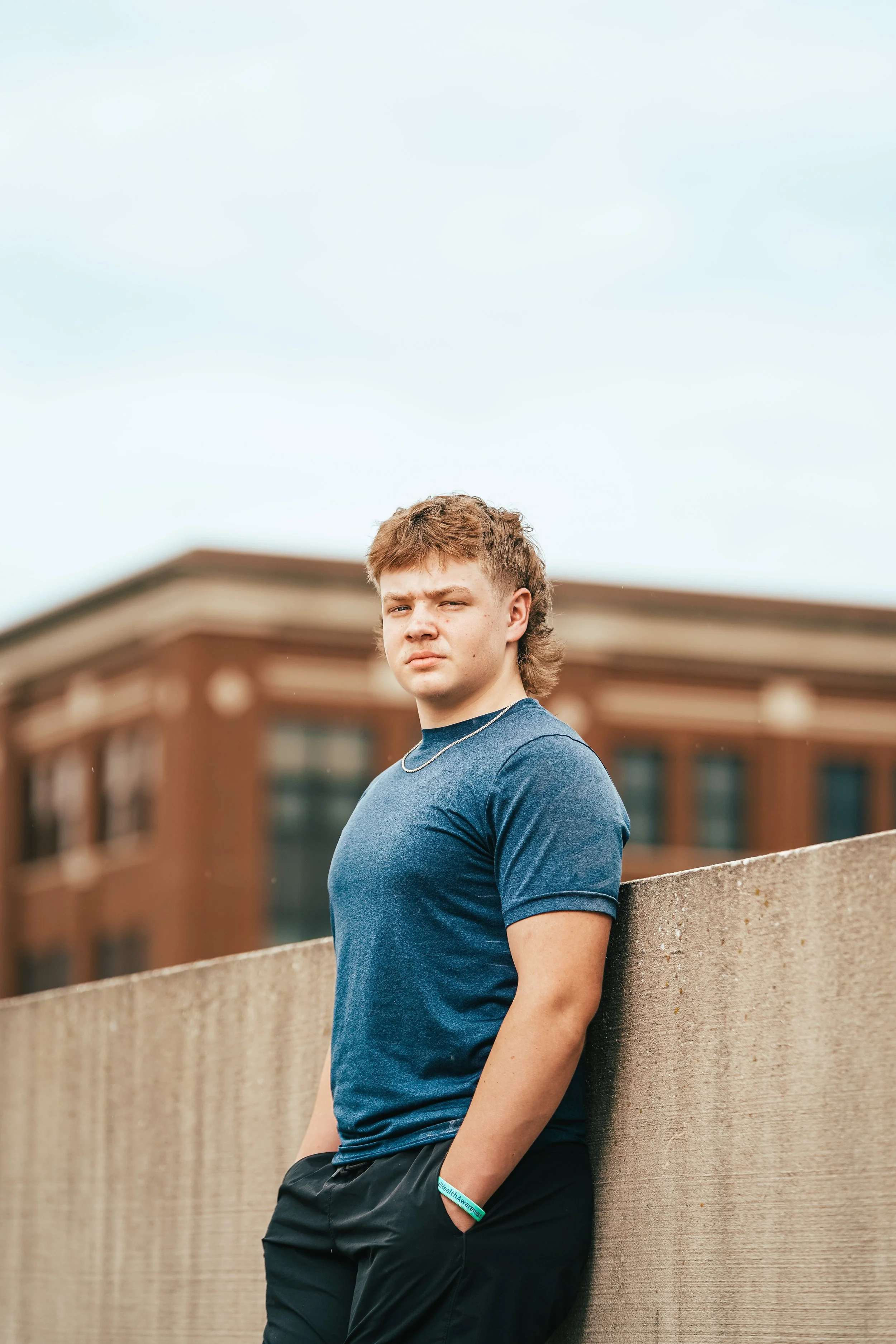 A young man with brown hair and a serious expression leaning against a concrete wall outdoors, with a blurred brick building in the background.