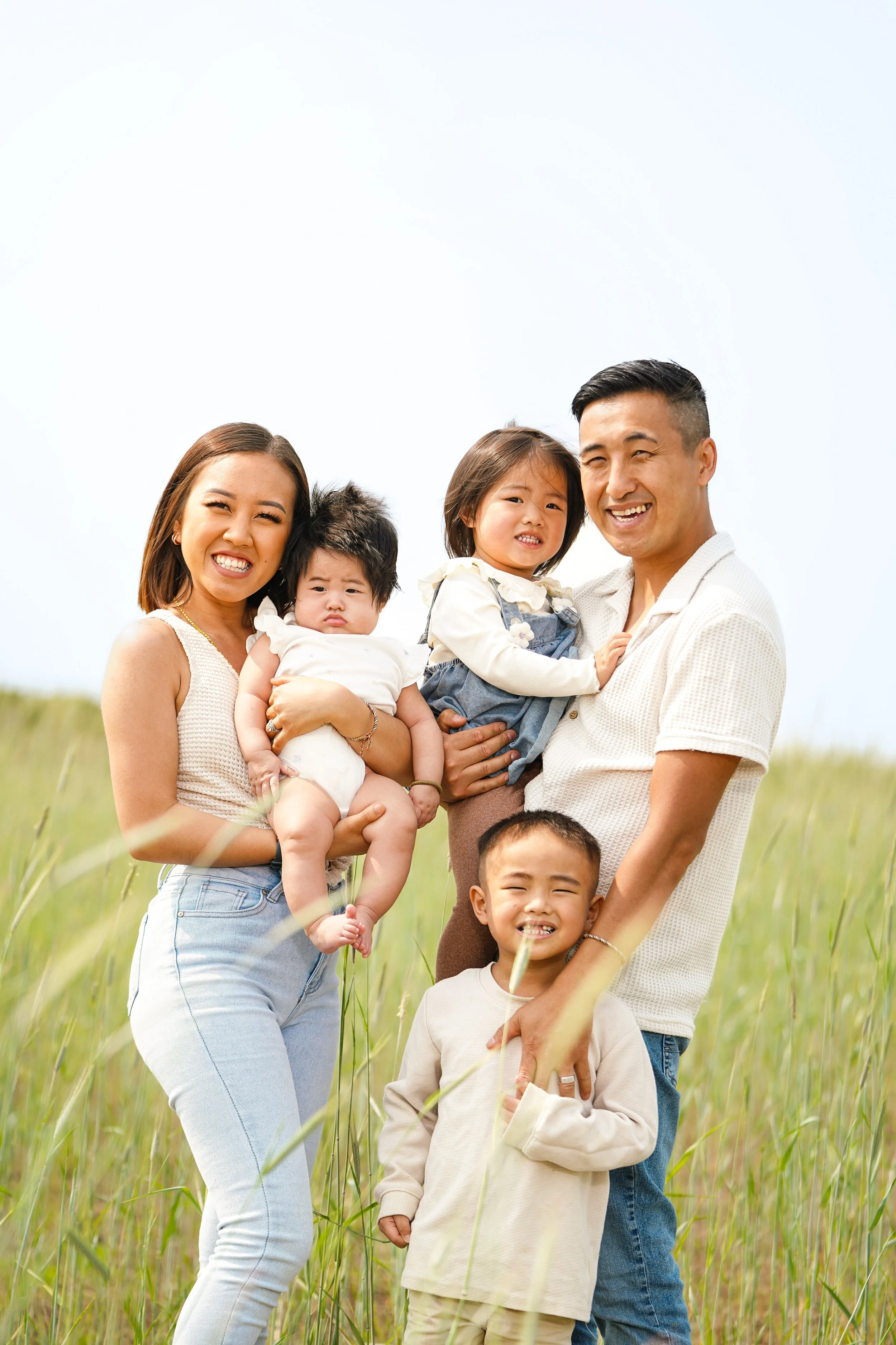 A multiethnic family of six smiling and posing together outdoors in a grassy field on a sunny day, including two women, two men, and two children.