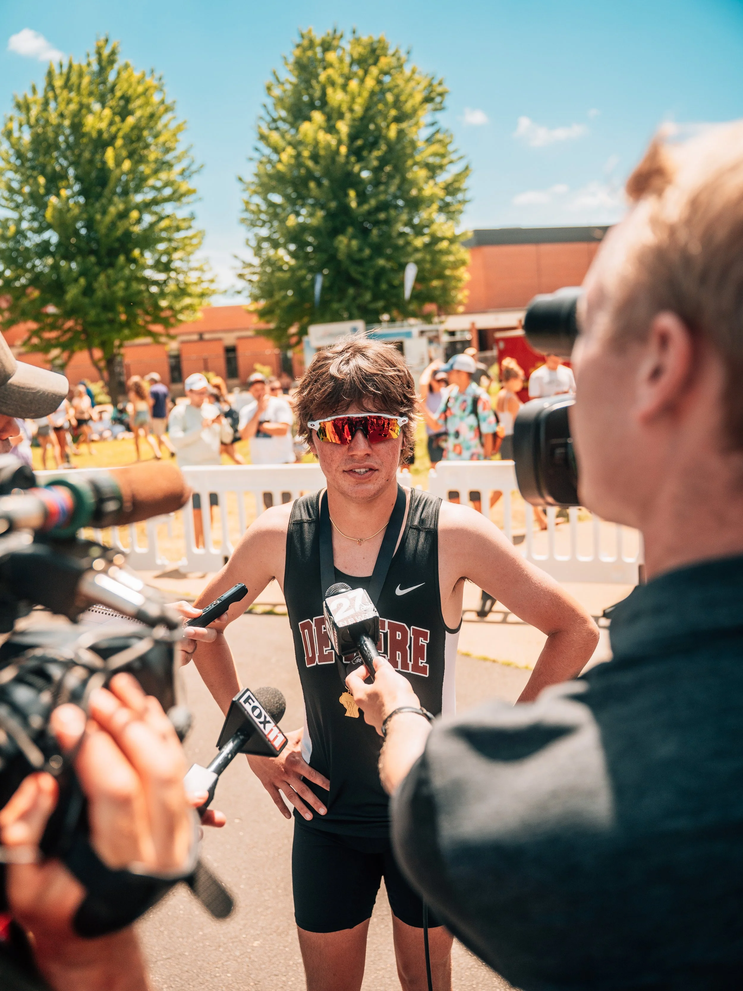 A young woman wearing sunglasses and a black athletic outfit being interviewed by multiple reporters holding microphones.