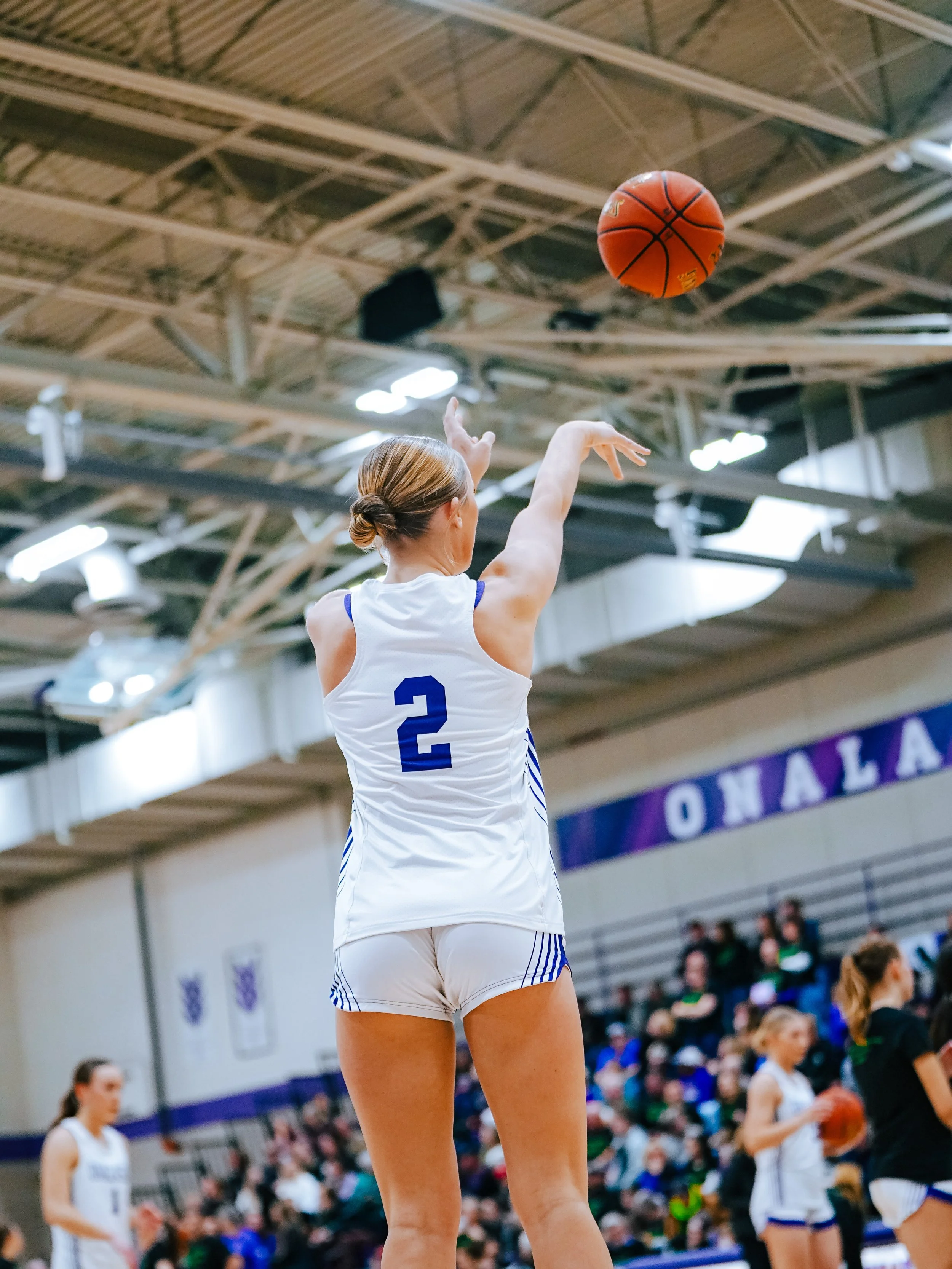 A female volleyball player in a white jersey with the number 2 is about to serve the ball during a match in a gymnasium. The gymnasium has a high ceiling with exposed metal beams and is filled with spectators.