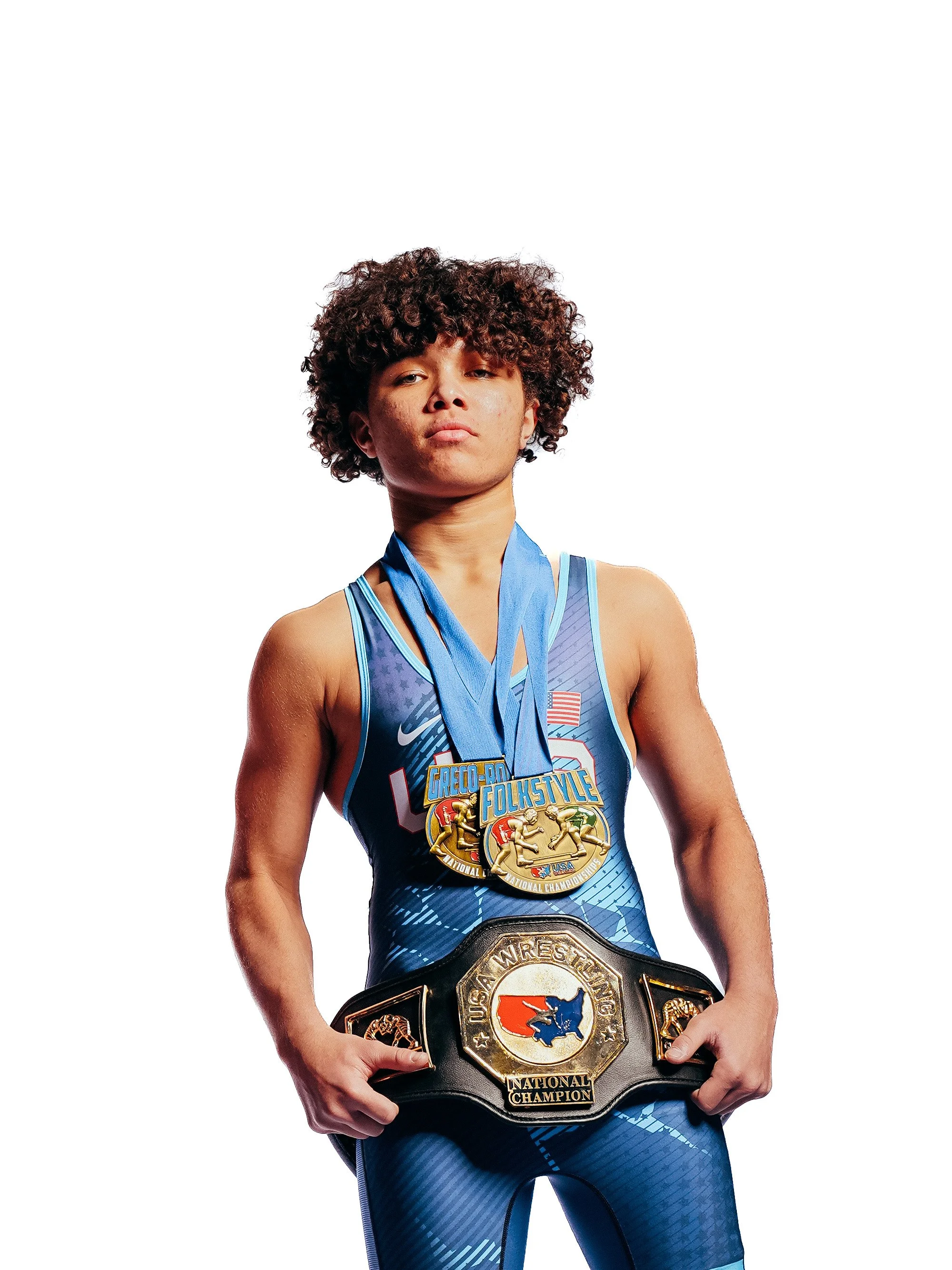 A female wrestler with curly hair wearing a blue athletic uniform, holding a championship belt, and wearing medals around her neck, against a plain white background.