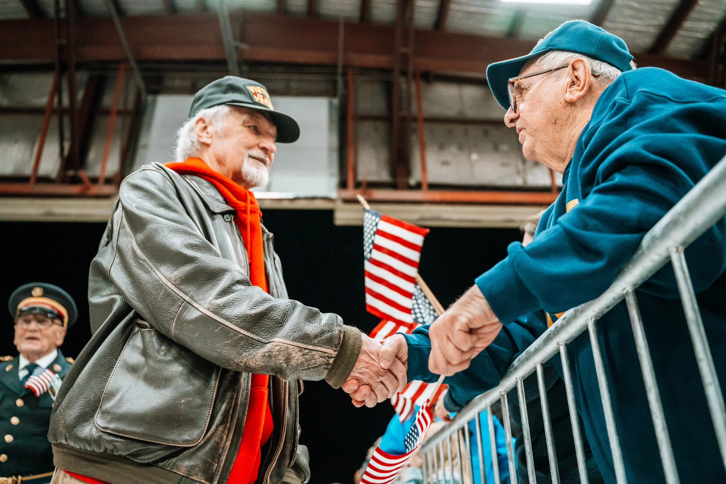 Two elderly men shake hands at a patriotic event, with American flags visible in the background. One man wears a green hat and leather jacket, the other wears a blue jacket and cap. A third man in a uniform stands behind them.