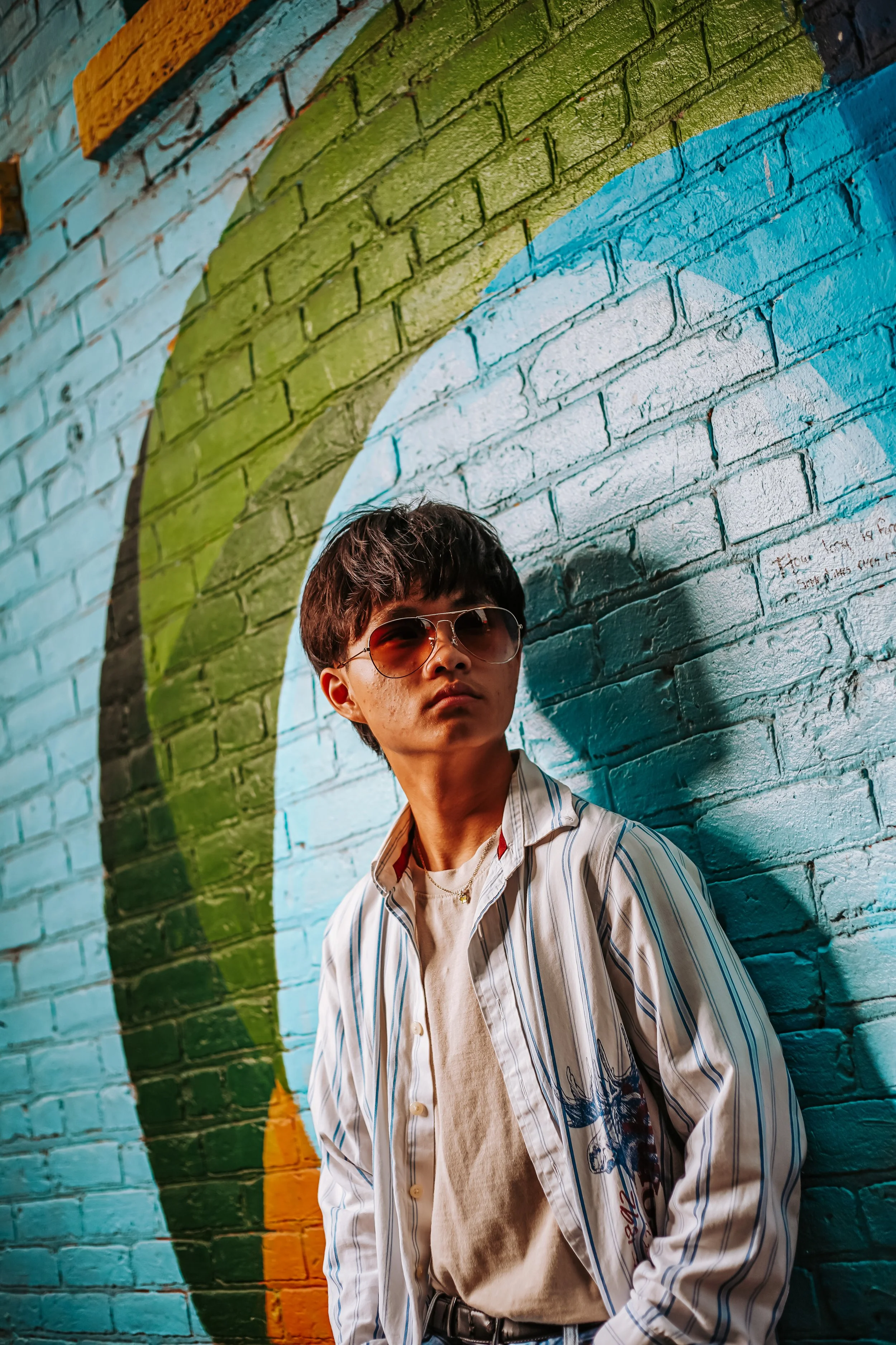 A young person with short brown hair wearing sunglasses, a white t-shirt, and a striped button-up shirt, standing against a colorful mural on a brick wall.