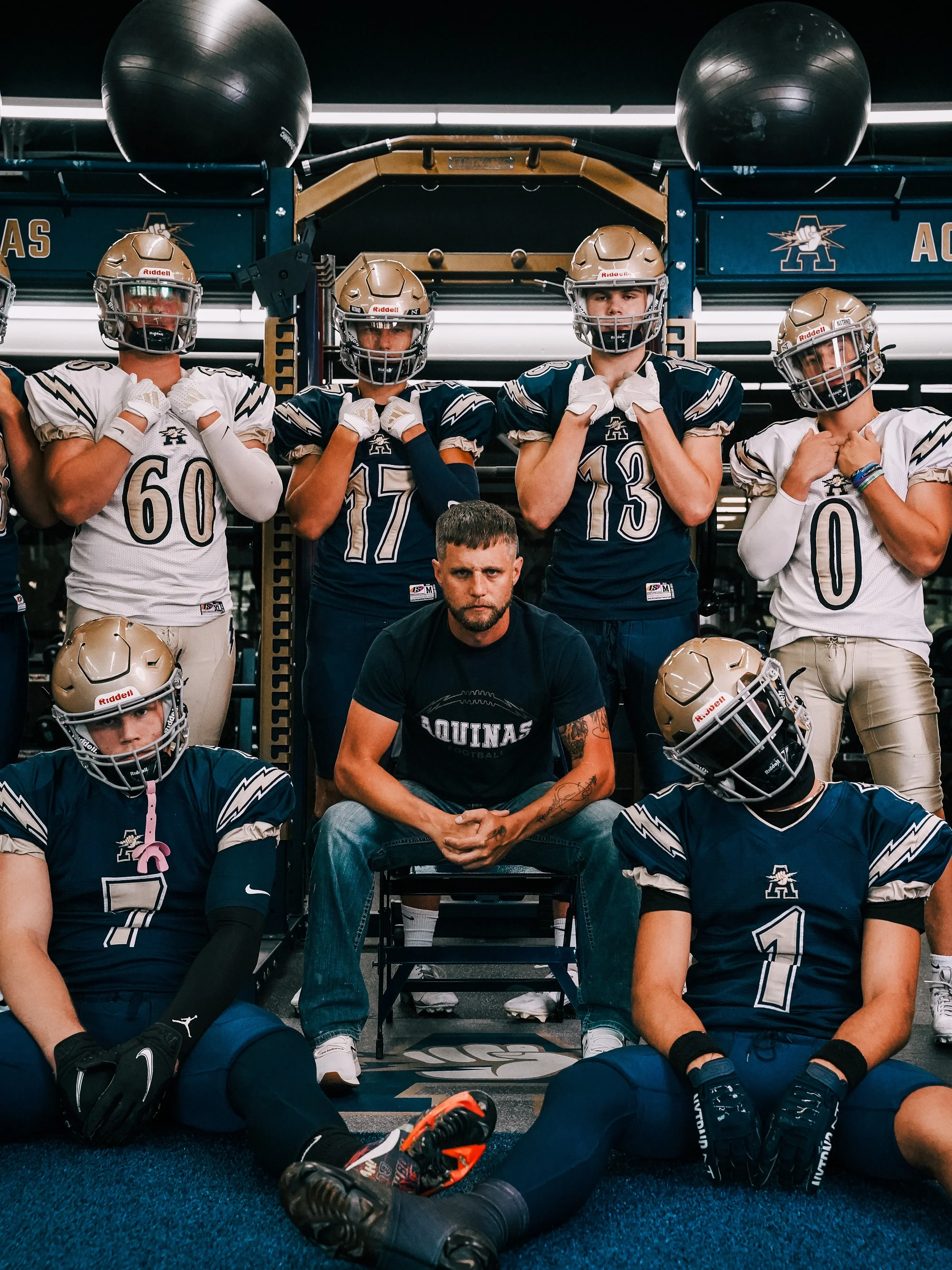 Football team members in gear sitting and standing in locker room with coach in center, all with hands over chest or face, some sitting on ground, wearing uniforms and helmets.