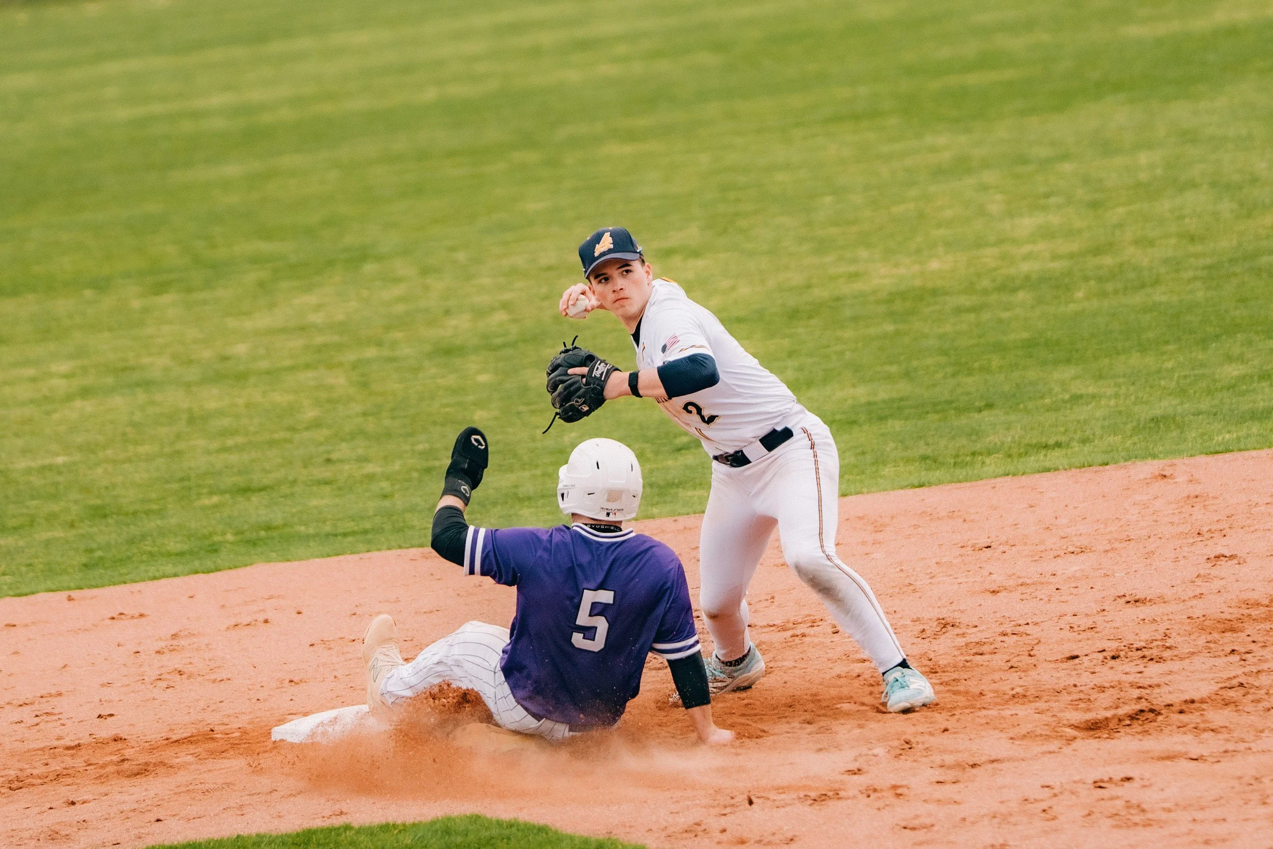 A baseball player in a white uniform with a navy cap and the number 2 on his jersey, looking alert and ready to catch the ball, as he stands on the field. A player in a purple uniform with the number 5 is sliding into a base nearby, with the baseman 