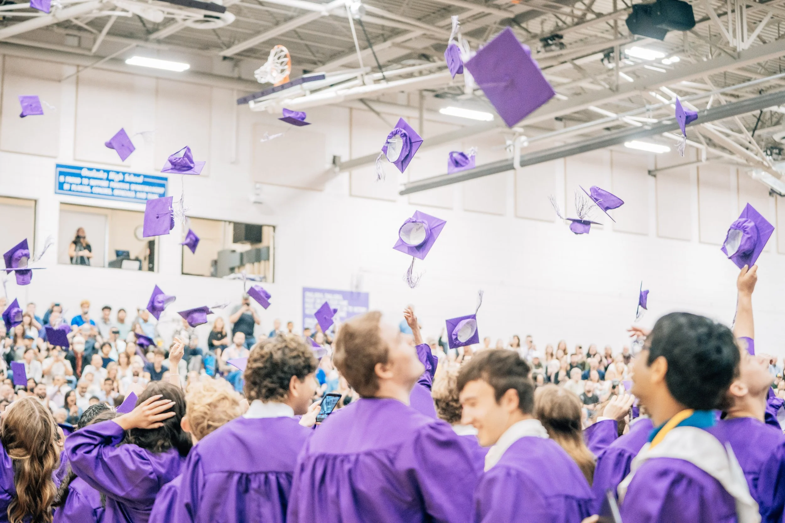A large indoor graduation ceremony with students in purple caps and gowns tossing their caps into the air, a packed audience seated in the background, and a digital screen on the wall.