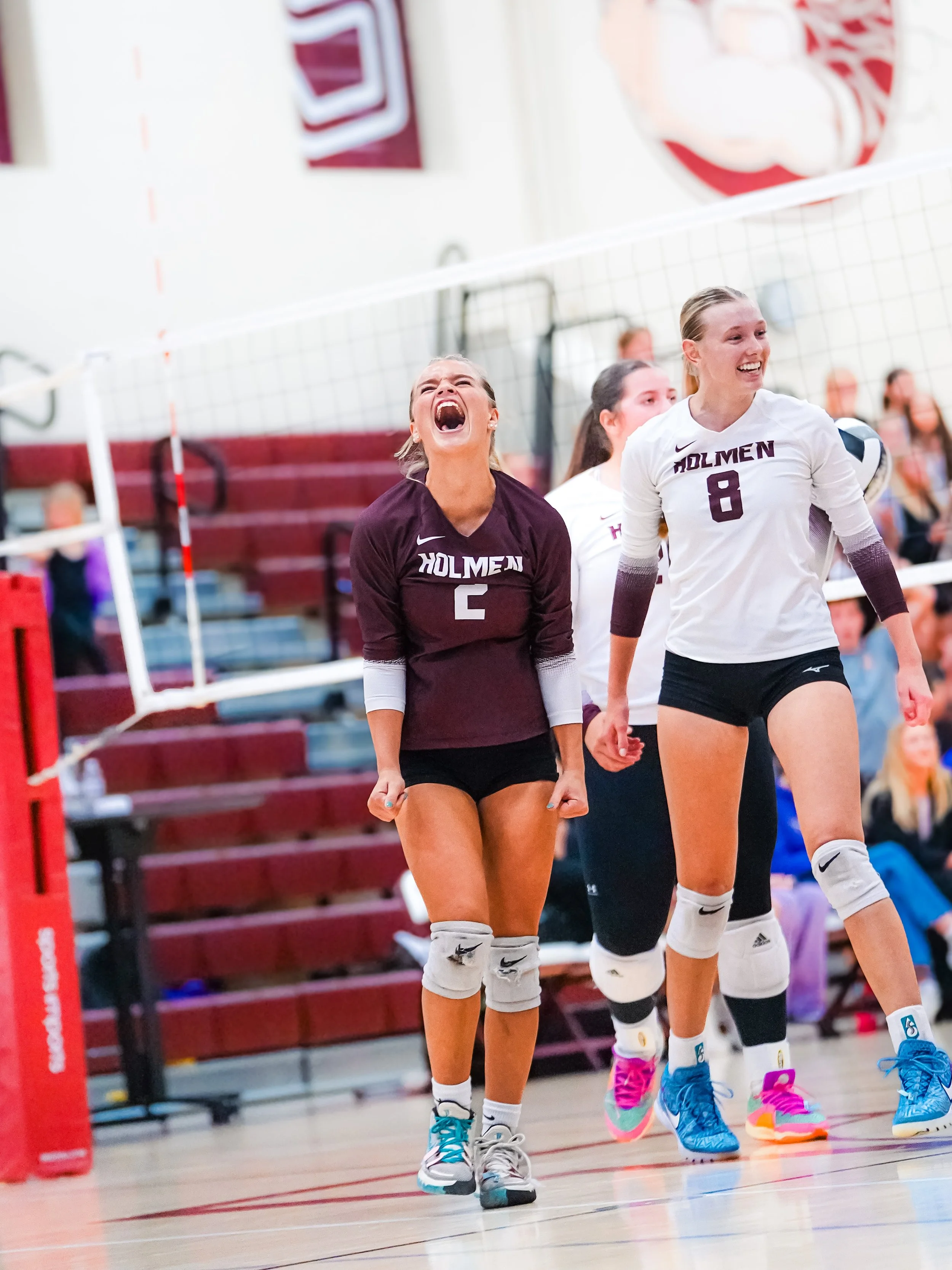 Female volleyball players celebrating on an indoor court, wearing white and maroon jerseys with the name "Holmen" and their numbers. They are smiling and appear joyful.